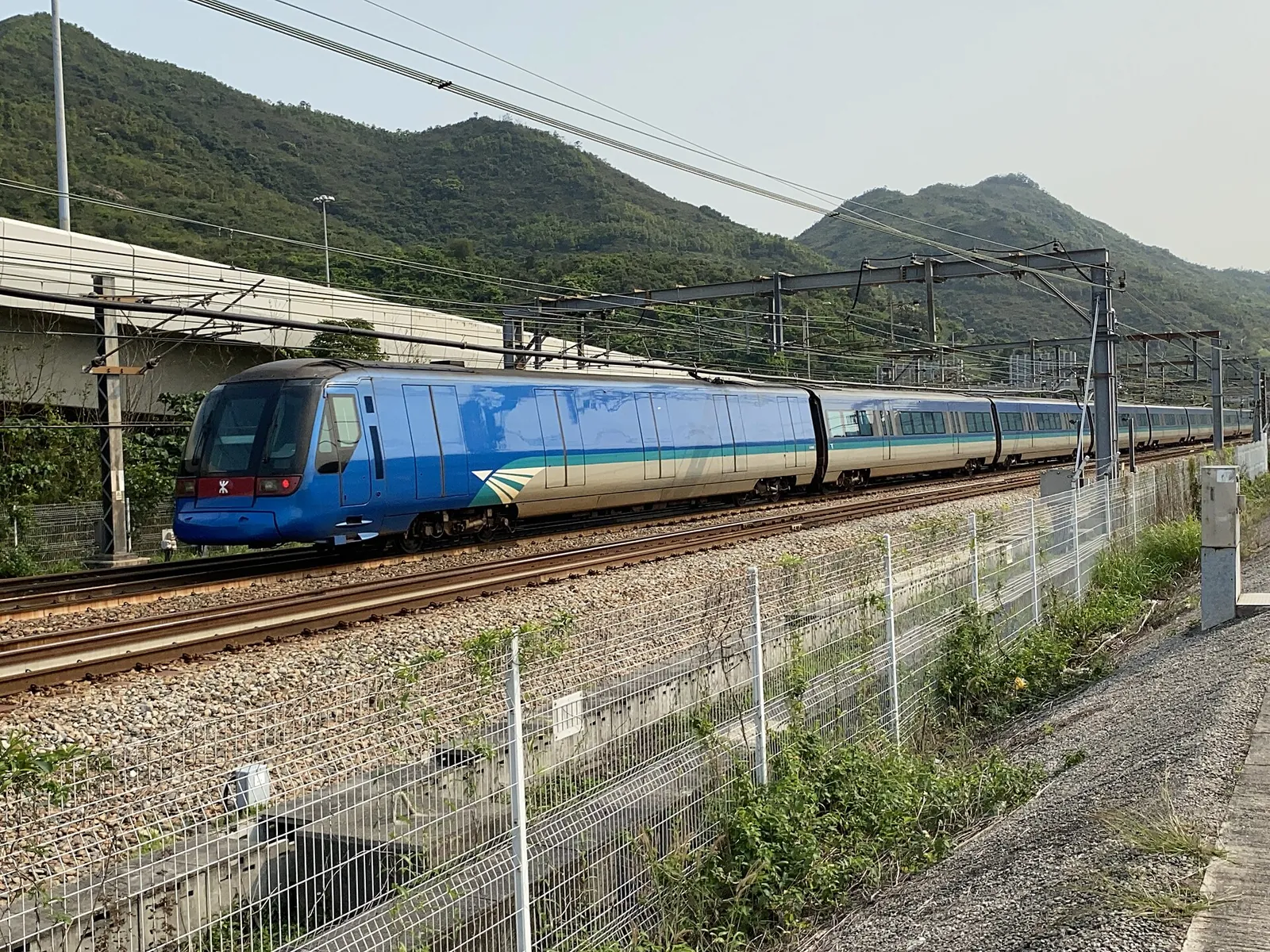 Modrý vlak Airport Express u zelených kopců v Sunny Bay