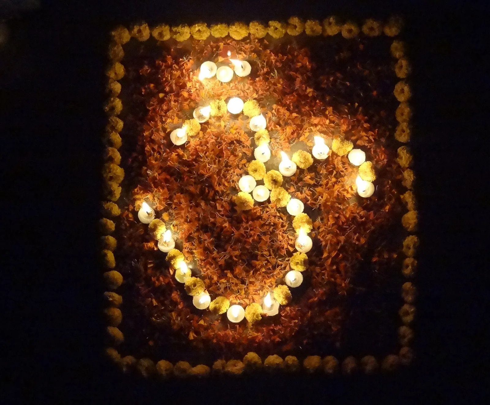 Symbol Óm vyložený z rozsvícených hliněných lamp a okvětních lístků aksamitníků na zemi v noci