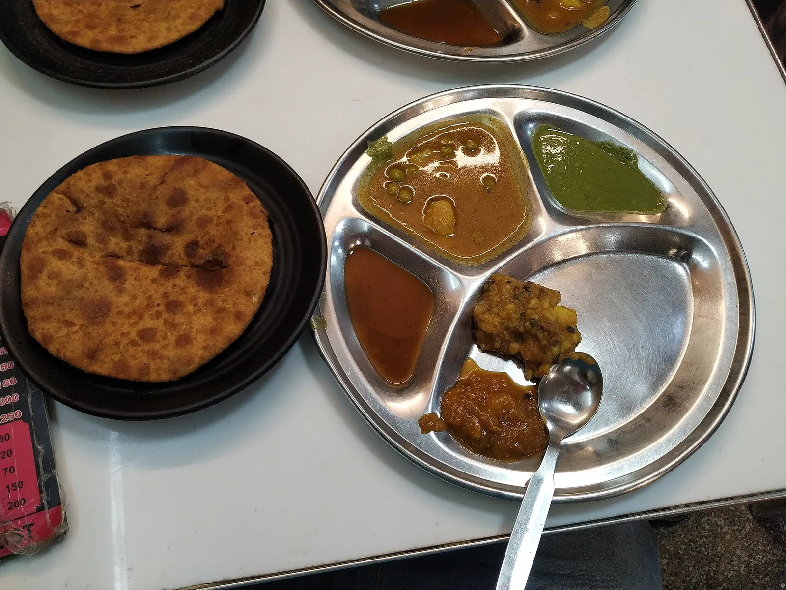 Ein Paratha auf einem schwarzen Teller neben einem Stahlteller mit Kartoffelcurry, Pickles und Chutneys in der Paranthe Wali Gali in Alt-Delhi