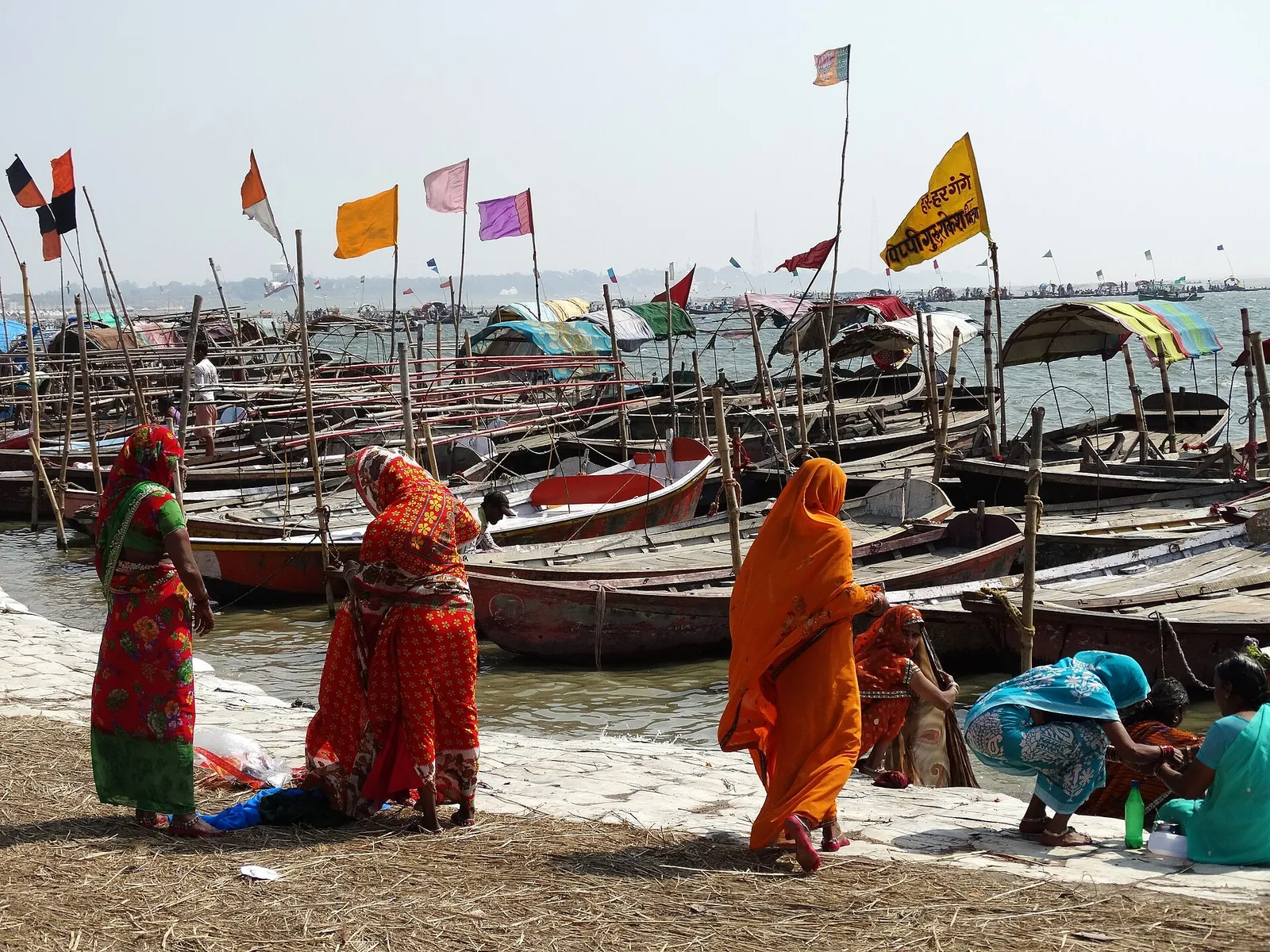 Mujeres con saris vivos en la orilla del río entre barcos y banderas durante la Magh Mela de Prayagraj