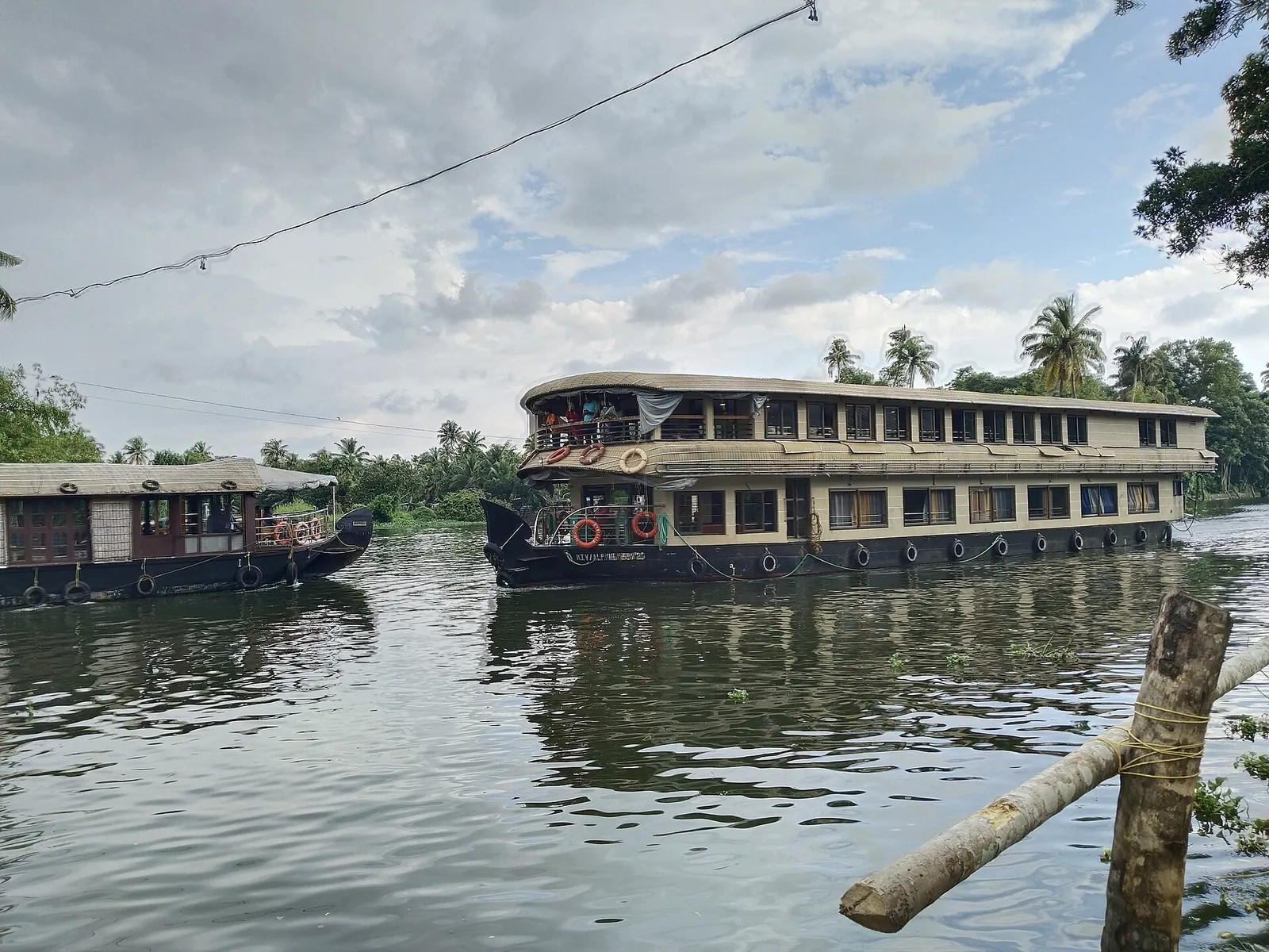 A large two-storey houseboat and a smaller one on Punnamada Lake under a cloudy sky, with palms on the far bank