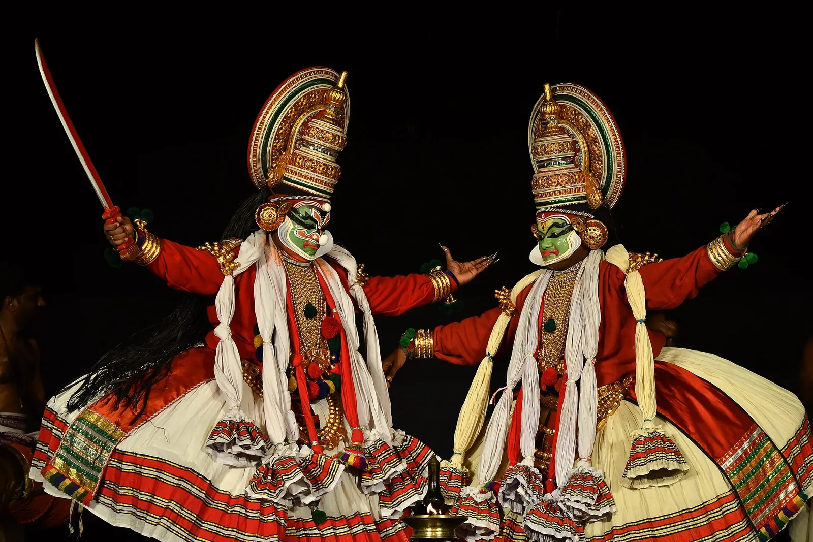 Doi interpreți de Kathakali cu machiaj pacha verde, podoabe de cap înalte și fuste largi roșii și albe, pe scenă