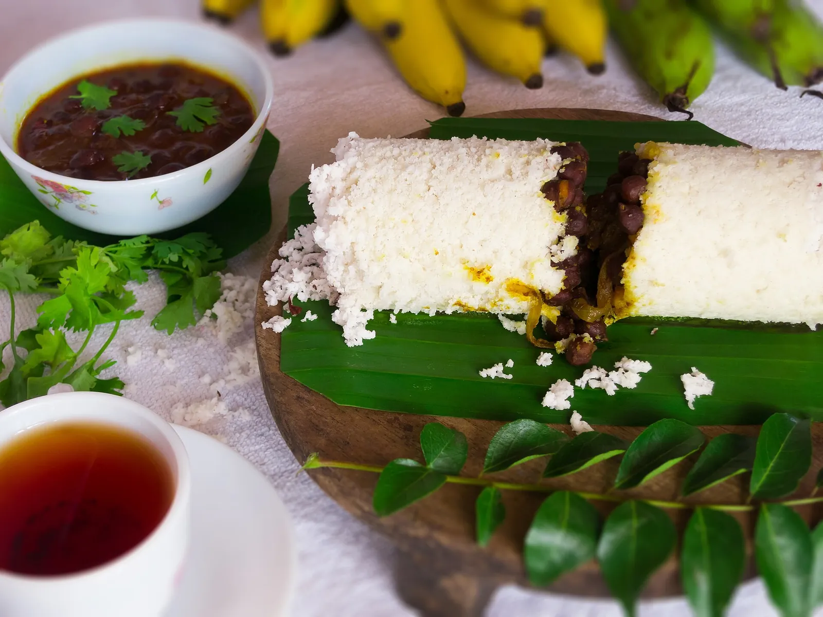 Doi cilindri de puttu alb pe o frunză de banan, cu un bol de curry închis la culoare de năut și o ceașcă de ceai
