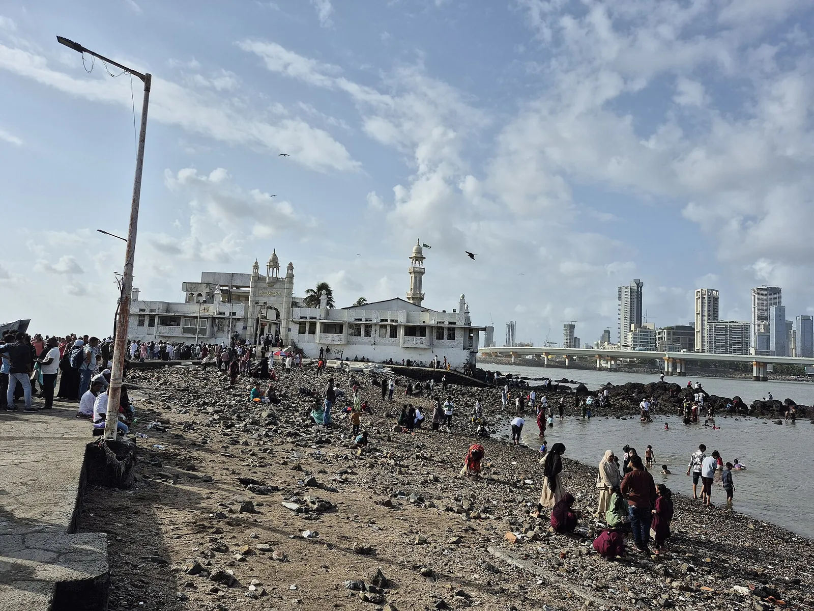 Haji Ali Dargah, cu cupolele albe și minaretul, în mare, în larg față de țărmul orașului Mumbai, cu vizitatori pe stânci