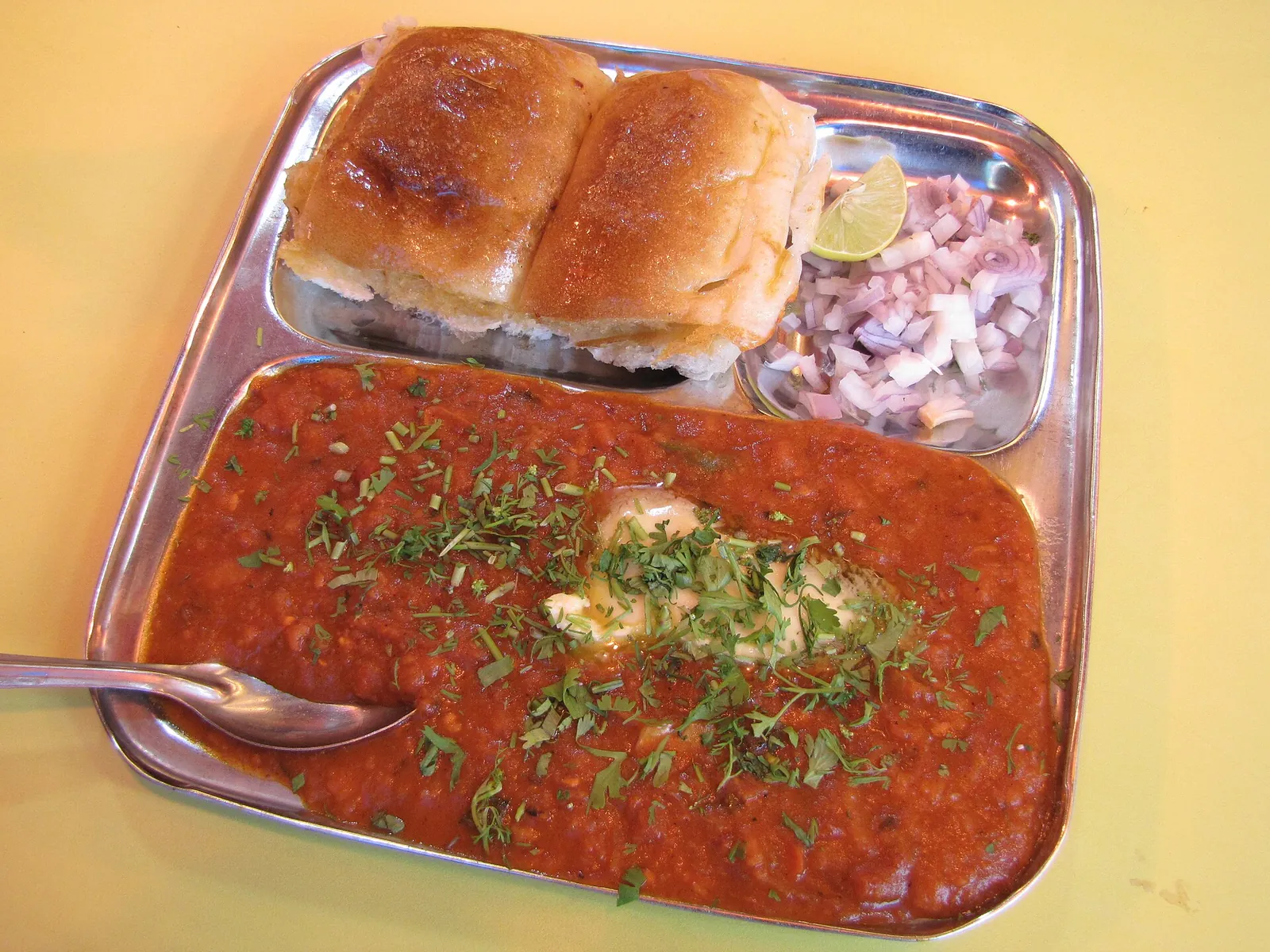 O farfurie de oțel cu pav bhaji: piure condimentat de legume cu unt și coriandru, două chifle unse cu unt, ceapă tocată și lime