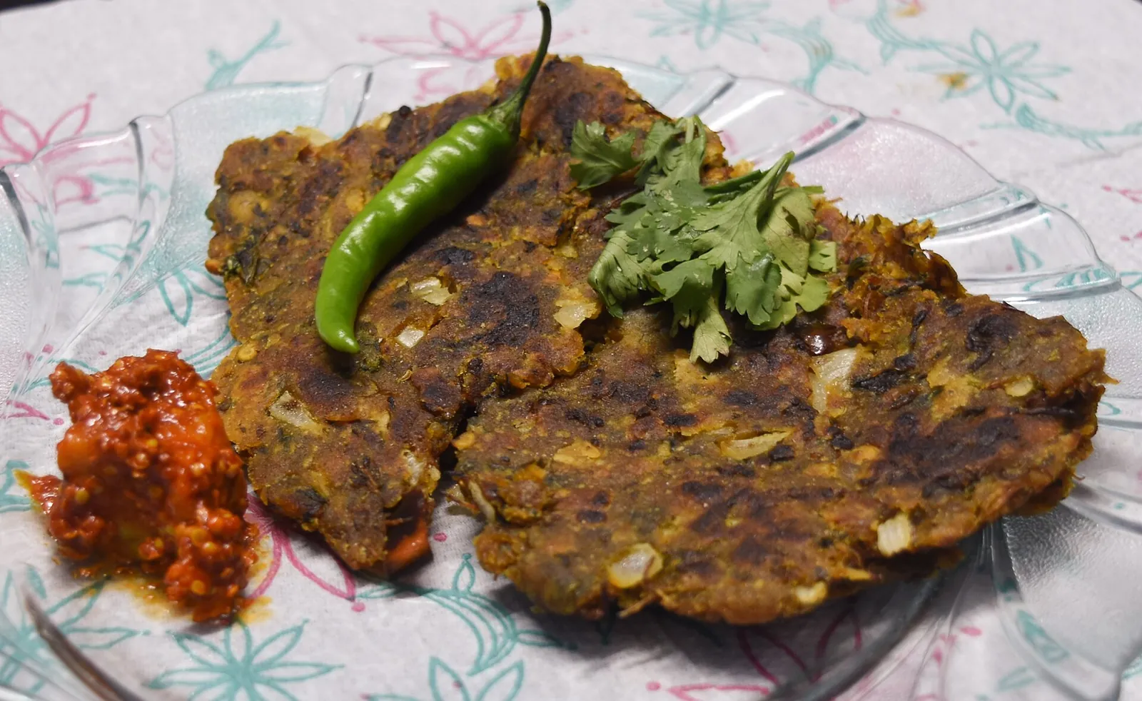 Dva ploché chleby thalipeeth s nakládanou přílohou, zelenou chilli papričkou a koriandrem na talíři