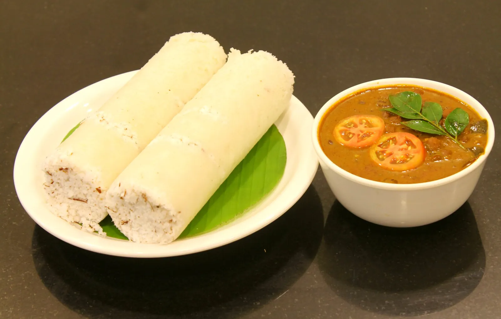 Dos cilindros de puttu al vapor sobre una hoja de plátano junto a un cuenco de curry de garbanzos