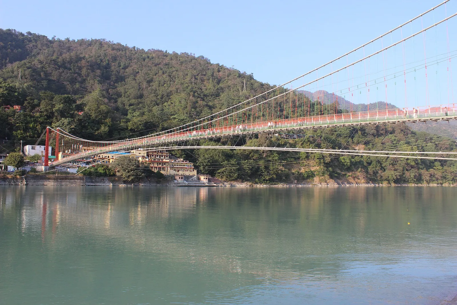 Die Hängebrücke Lakshman Jhula über dem grünen Ganga bei Rishikesh, mit bewaldeten Hügeln dahinter
