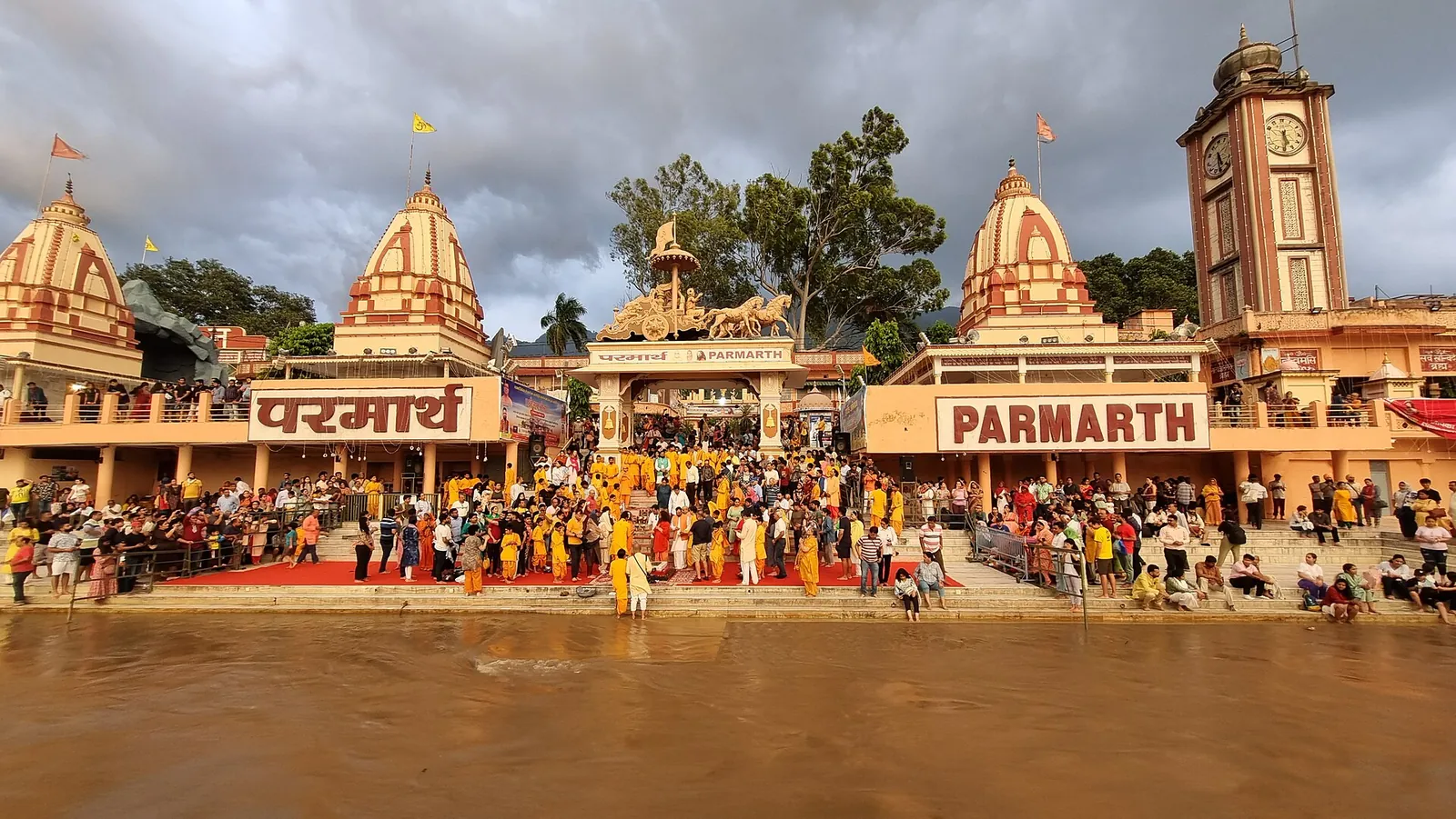 Menschenmengen auf den Stufen des Ghats von Parmarth Niketan am Ganga in Rishikesh, unterhalb der Tempeltürme des Ashrams