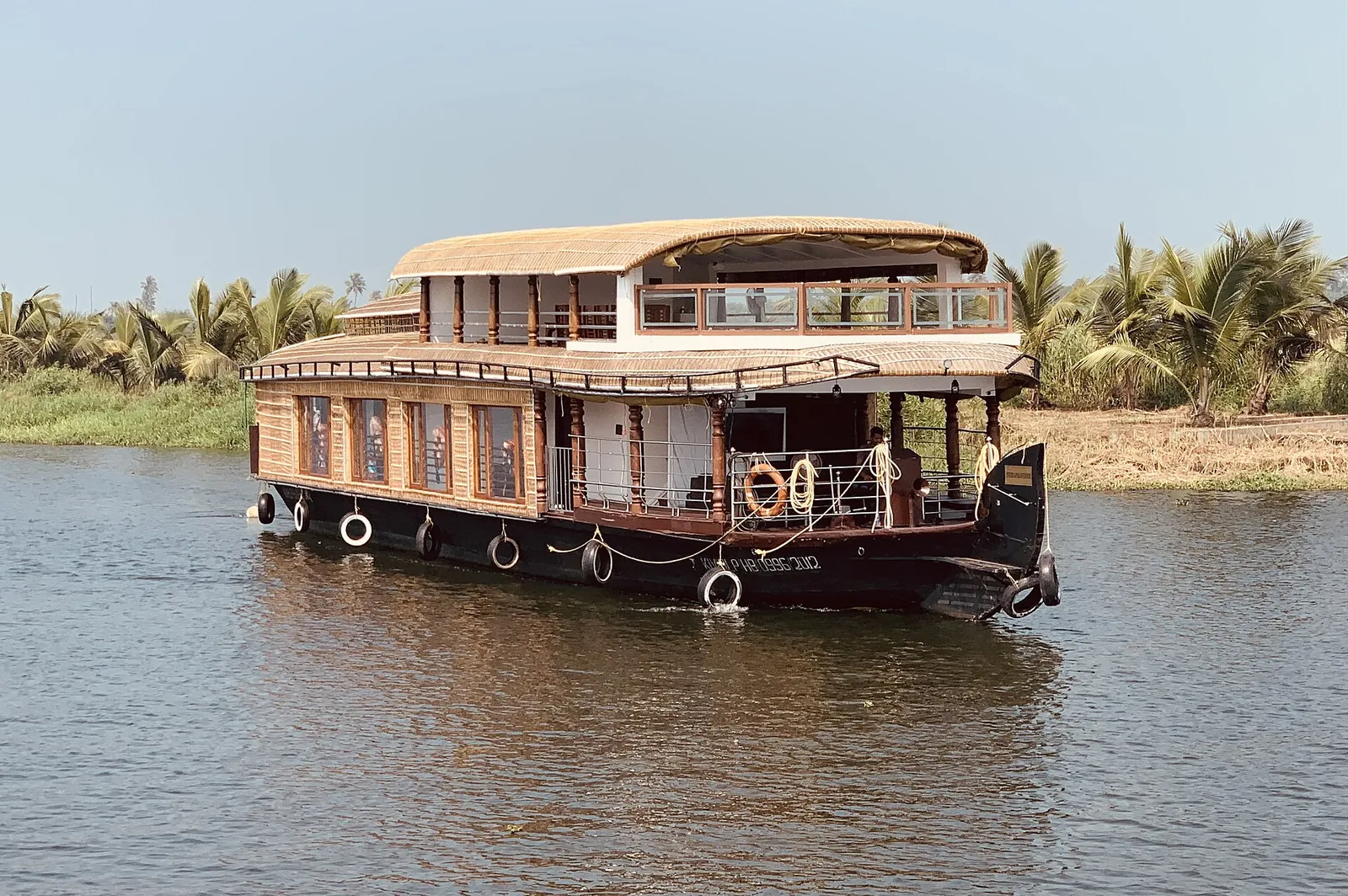 A two-storey wooden houseboat on the Alleppey backwaters, with palm trees on the bank