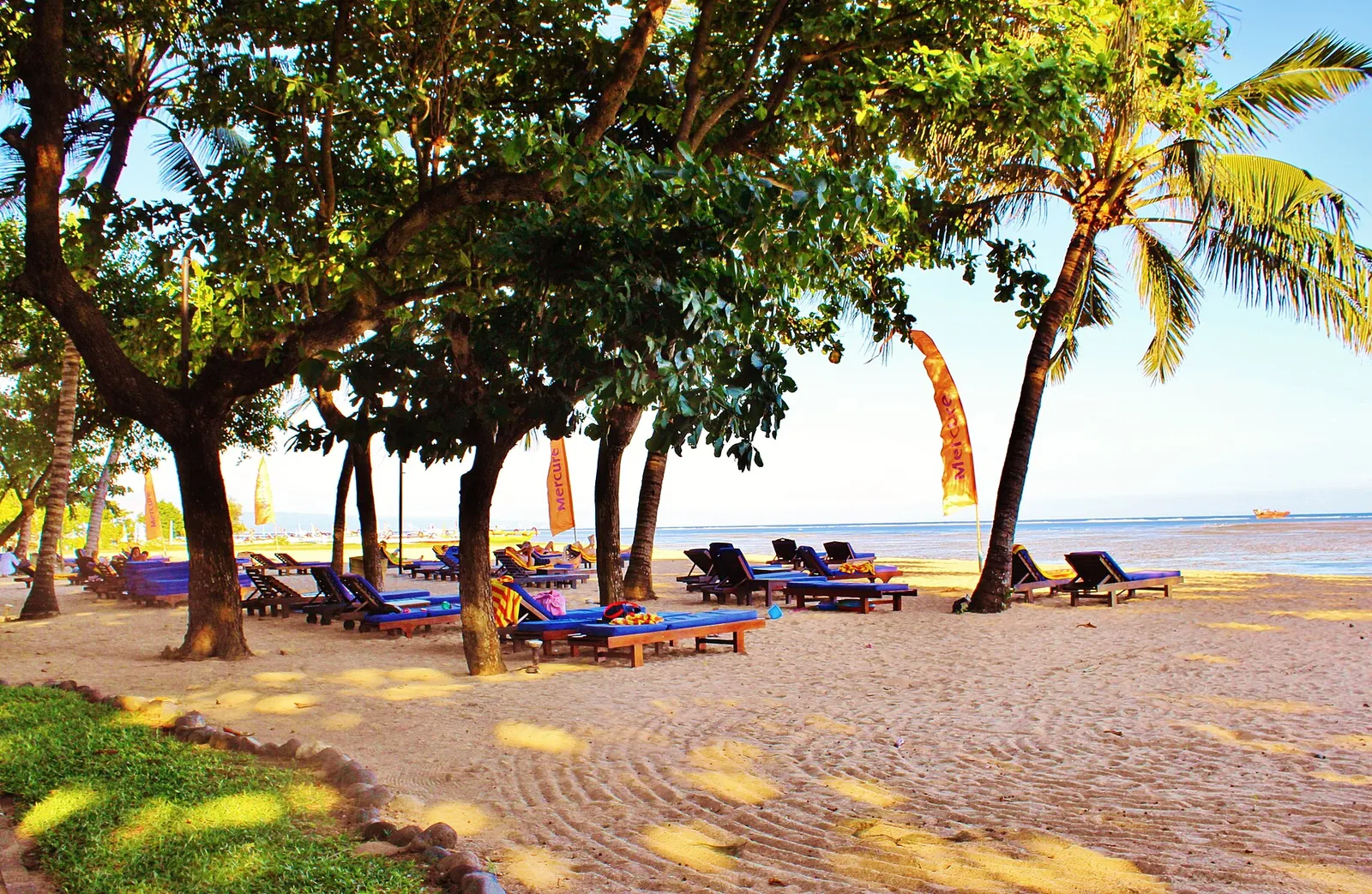 Sun loungers under shady trees on the sand at Sanur Beach, Bali, with the sea behind