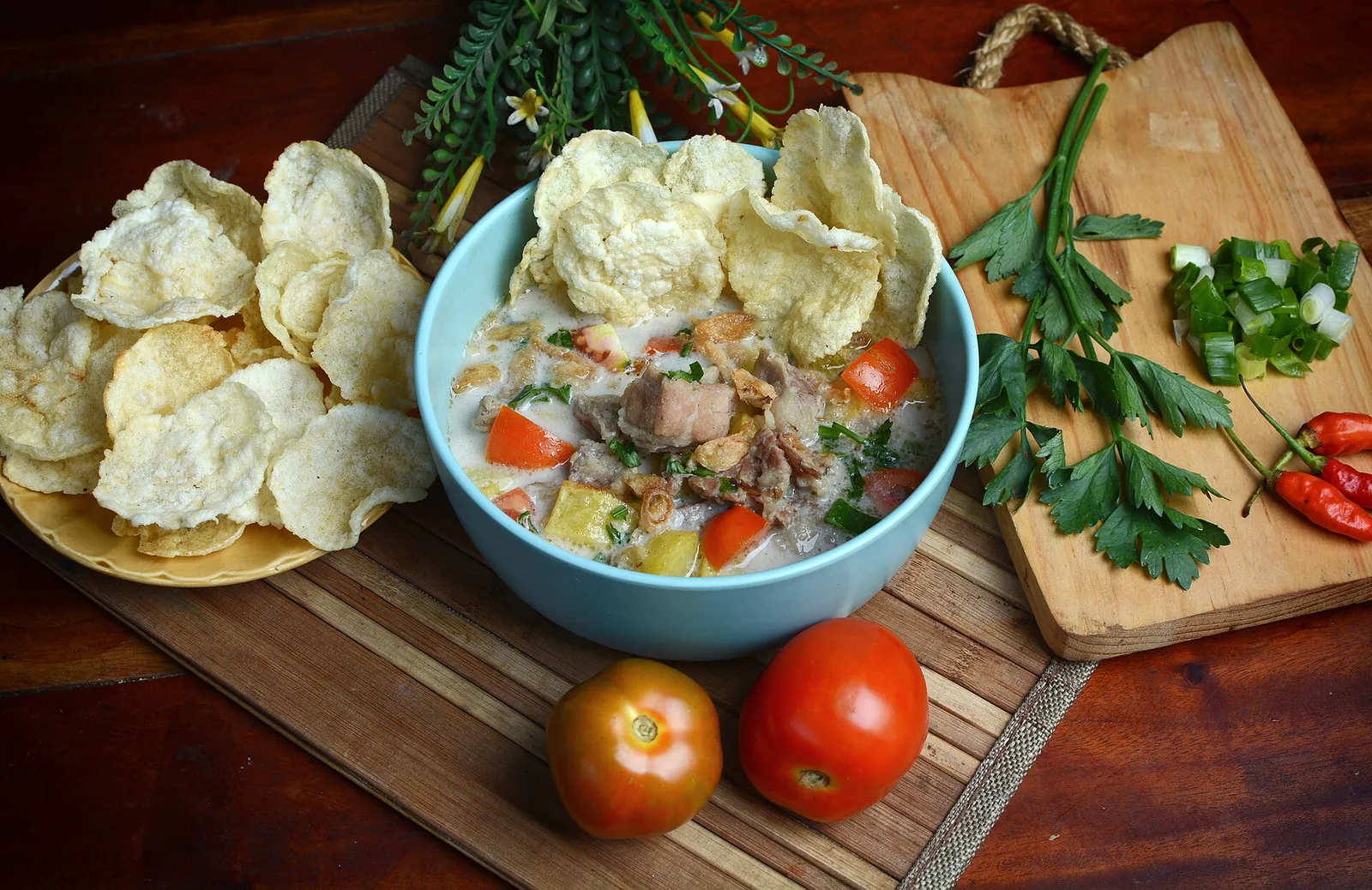 A bowl of soto Betawi with beef in a creamy coconut-milk broth, tomatoes and emping crackers on a wooden board