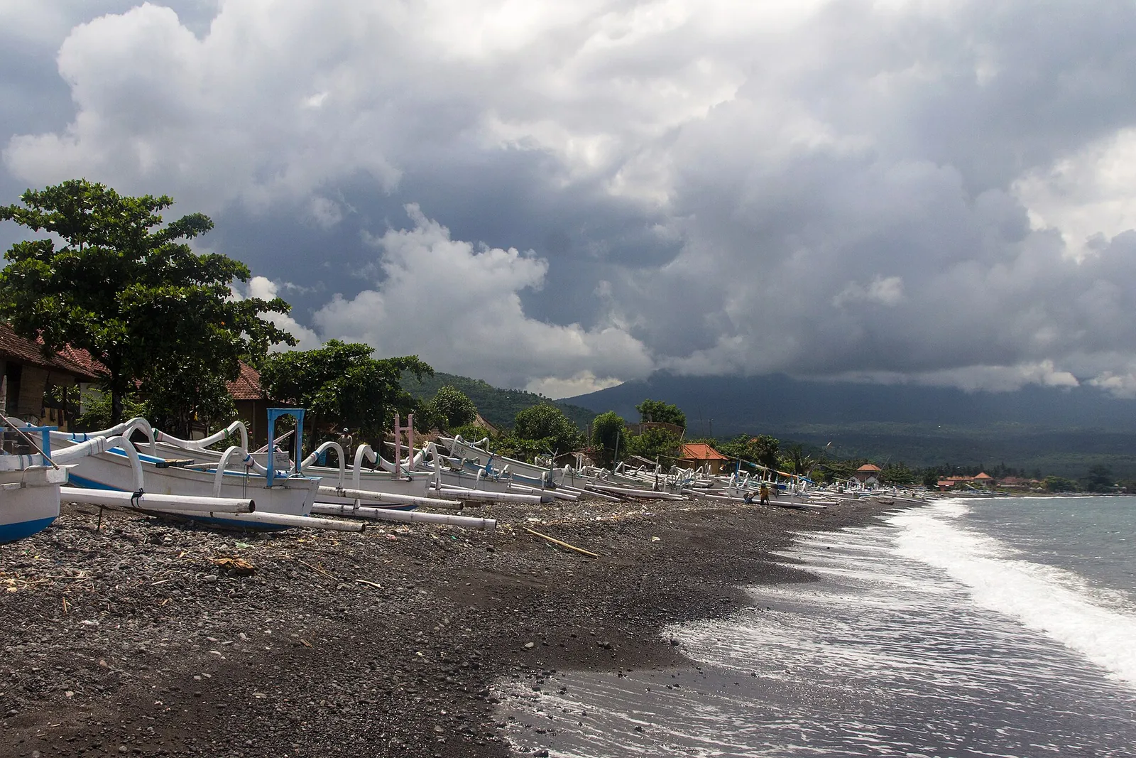 Black-sand beach at Amed lined with outrigger jukung boats under a cloudy sky