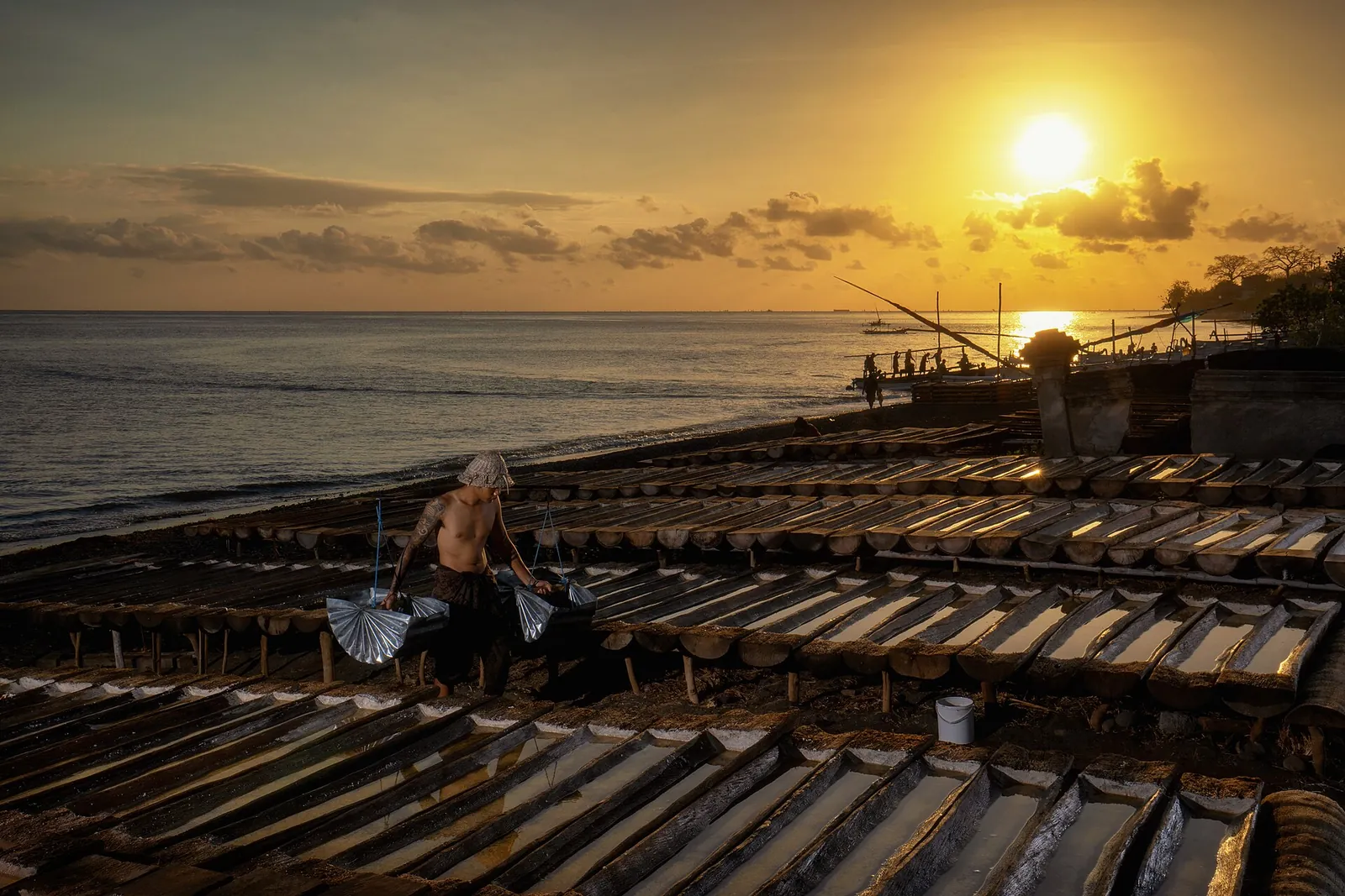 Salt maker carrying baskets past rows of wooden salt-drying troughs on Amed beach, with the sun low over the sea