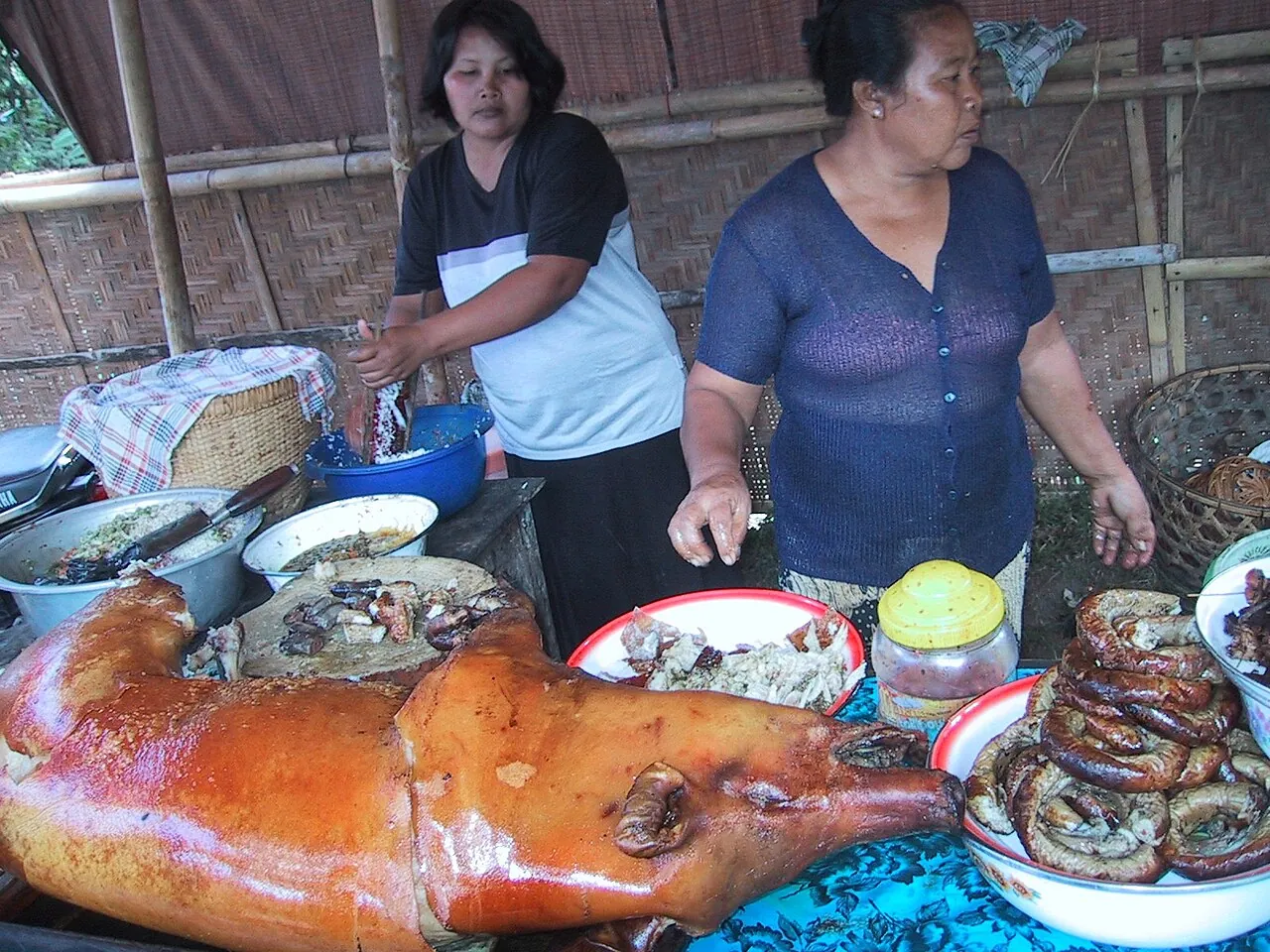 A whole roast suckling pig on a stall in Bali, with two women serving dishes beside it