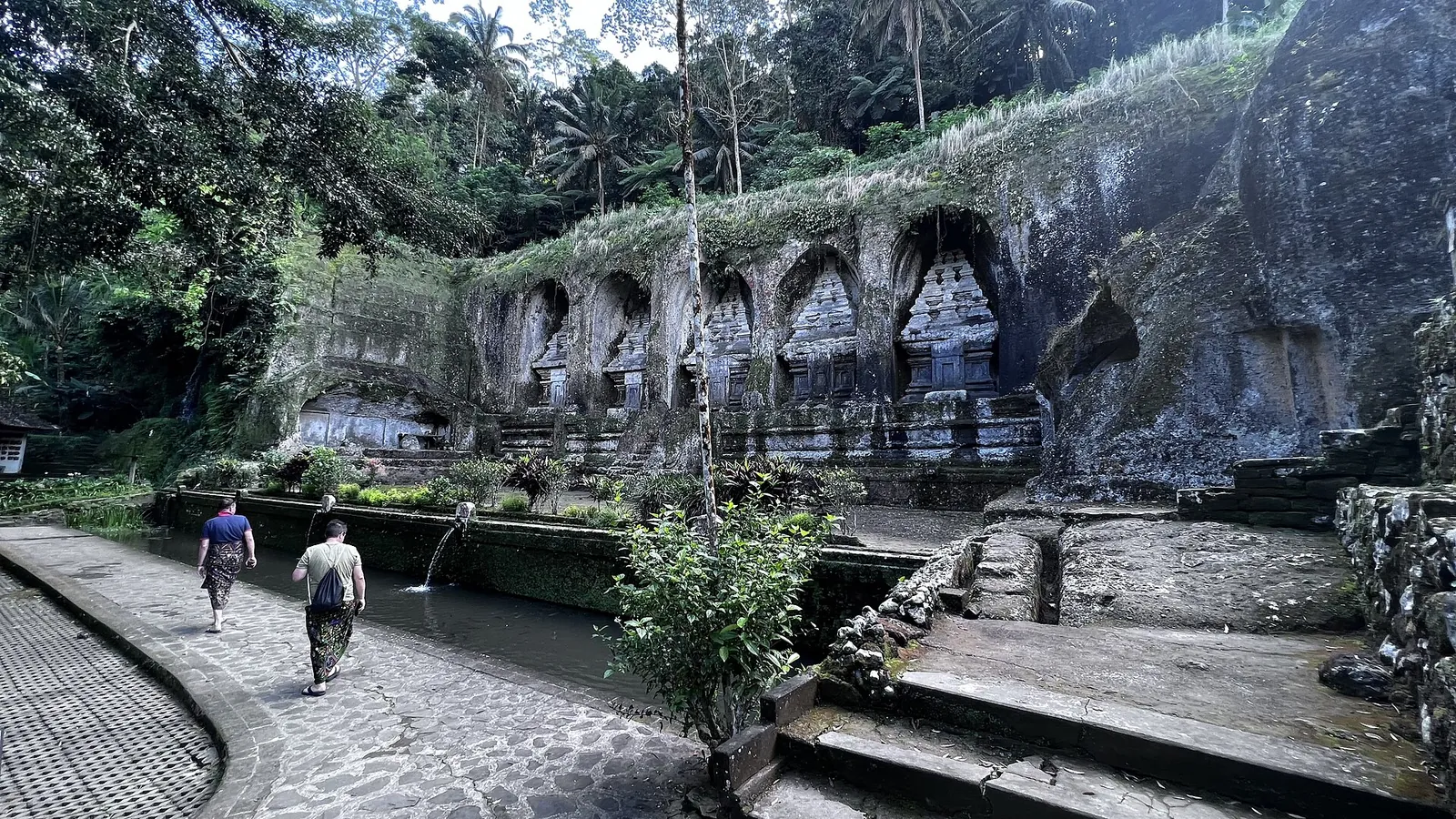 Shrines carved into the rock face at Gunung Kawi, with visitors walking along a paved path below