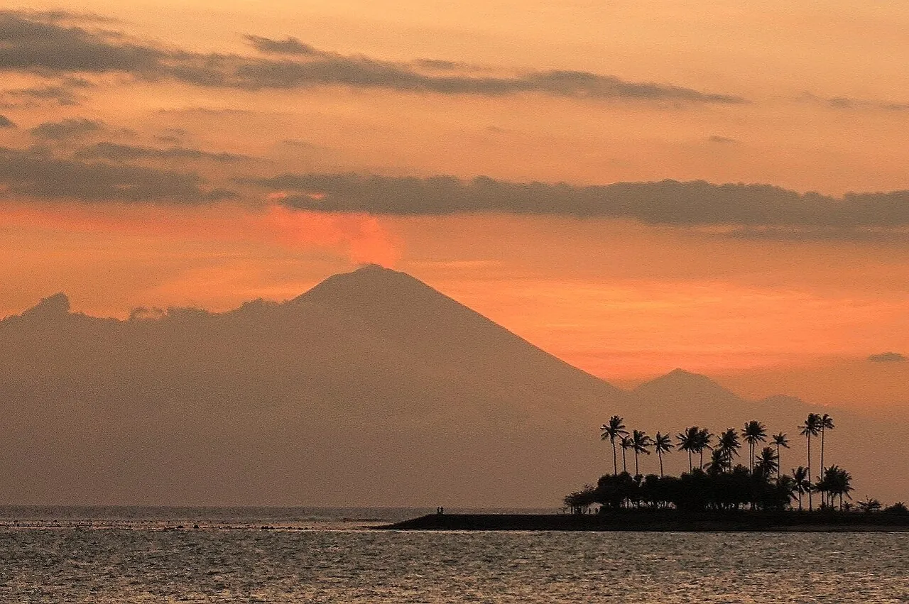 The cone of Mount Agung on Bali silhouetted against an orange sunset sky, seen across the Lombok Strait from Senggigi with palm trees on a spit of land