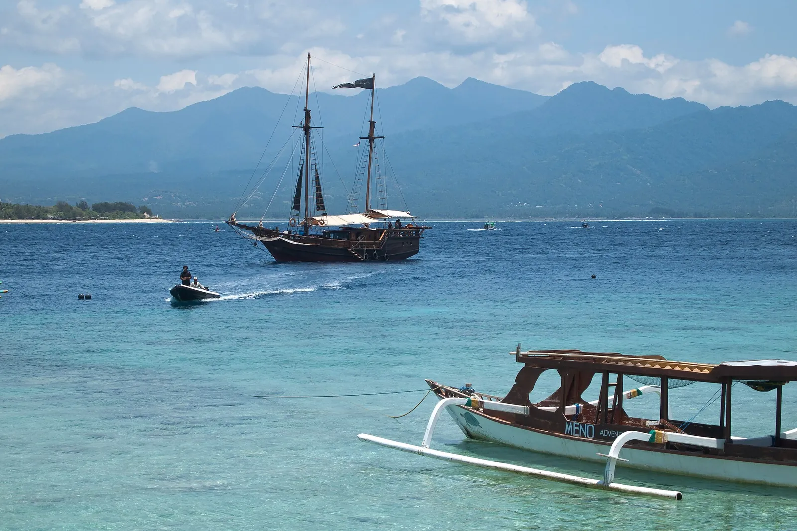 A two-masted sailing ship and a wooden outrigger boat on turquoise water off Gili Meno, with Lombok and the slopes of Mount Rinjani behind