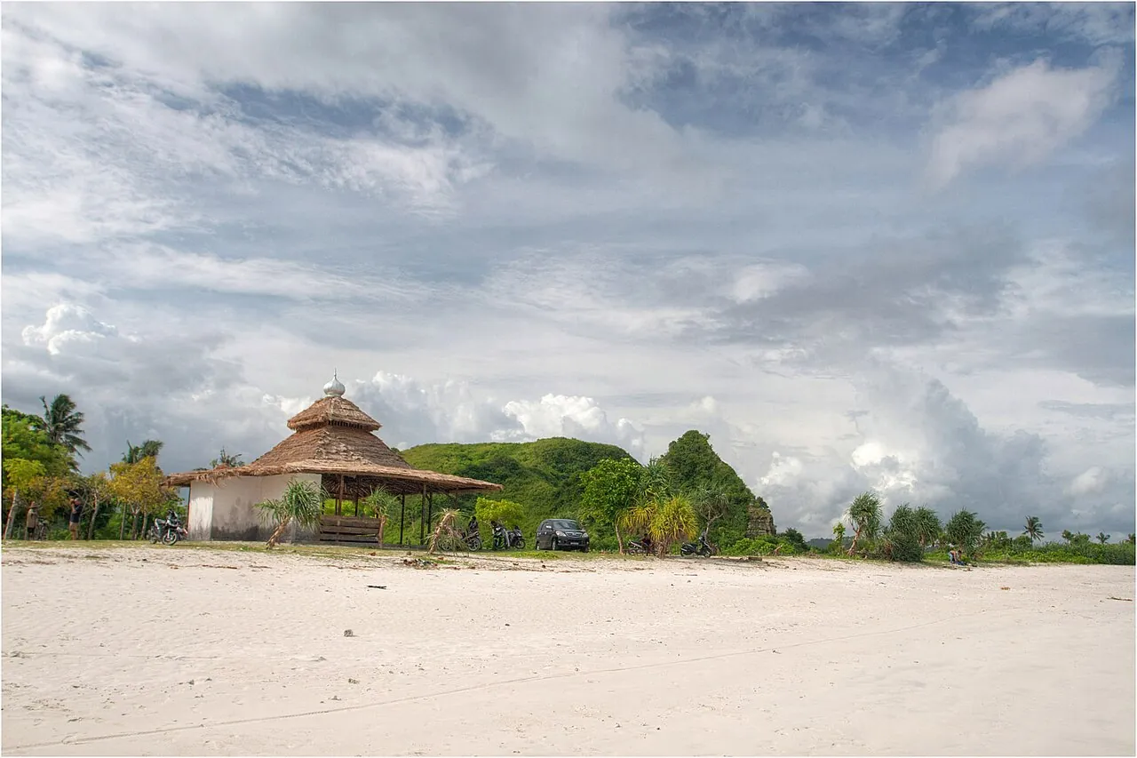 An empty white-sand beach with a thatched hut and green hills under a cloudy sky at Tanjung Aan, Lombok