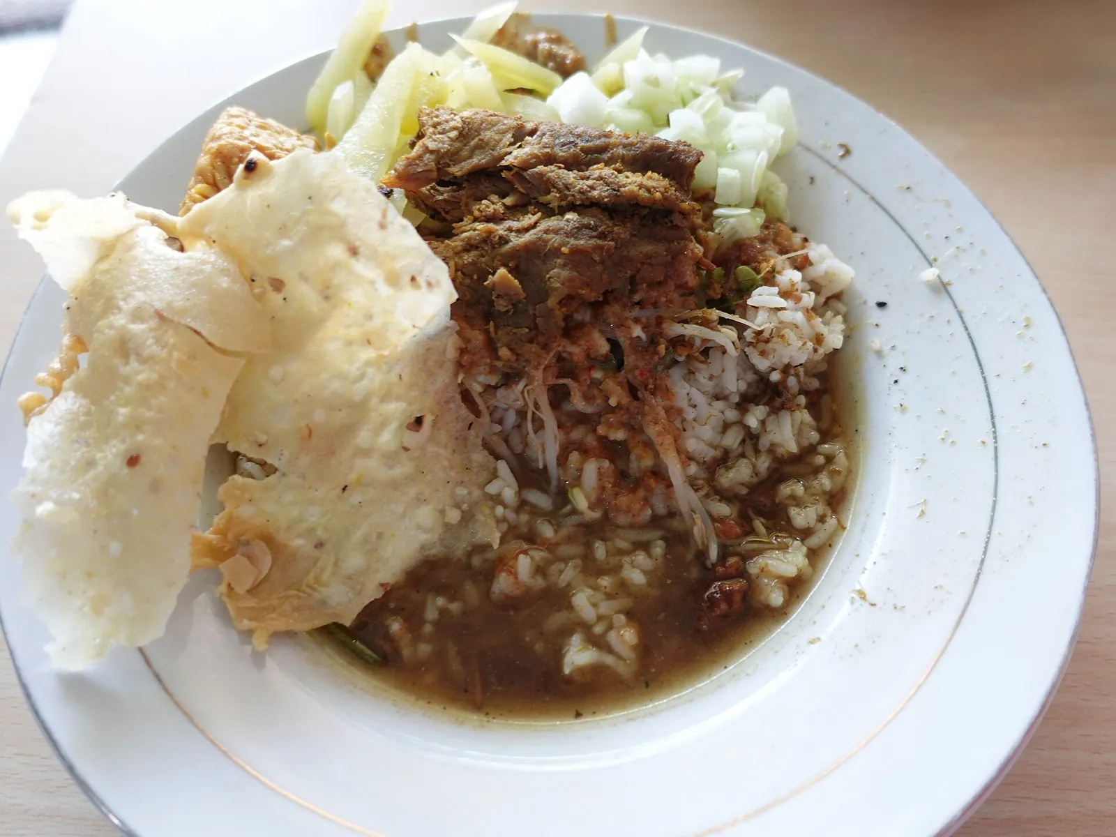 Rice with a slice of beef dendeng in dark rawon broth, vegetables, cucumber and rice crackers on a white plate