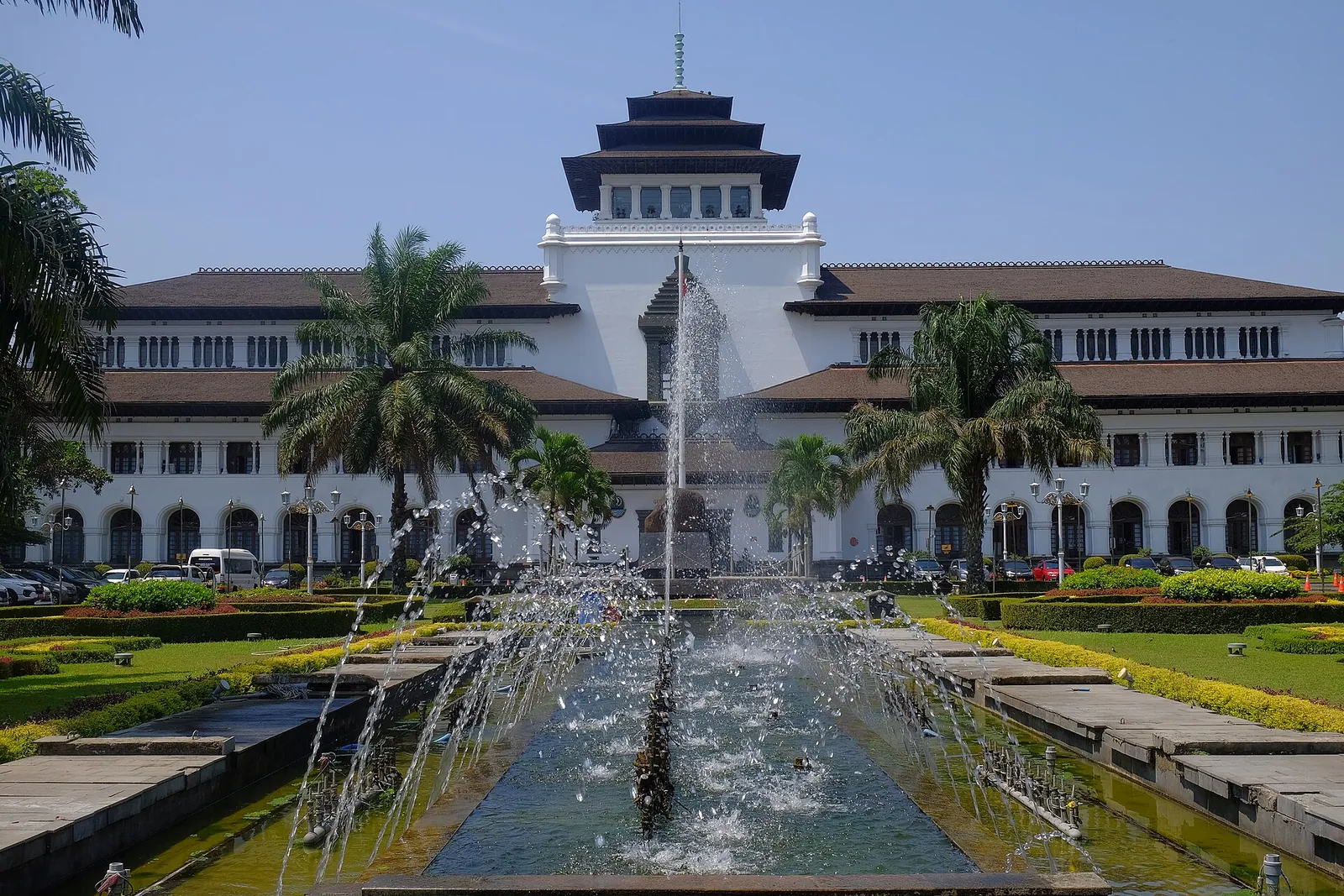 Gedung Sate in Bandung, a long white colonial building with a tiered central tower, behind a fountain and flower beds under a blue sky