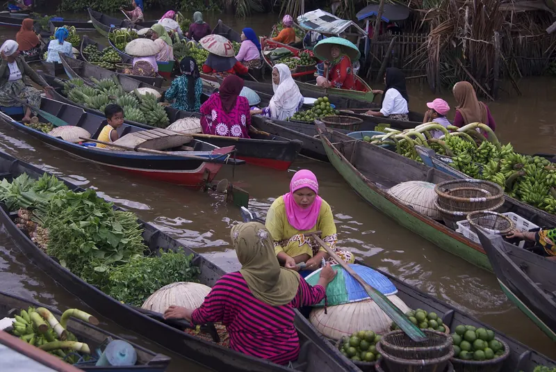 Women in headscarves and wide hats selling vegetables, fruit and greens from wooden boats at the Lok Baintan floating market