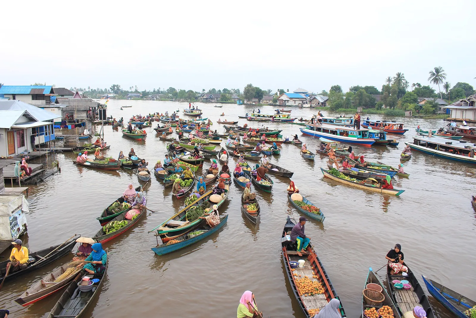 Dozens of small wooden boats crowding a wide brown river beside stilt houses at Lok Baintan