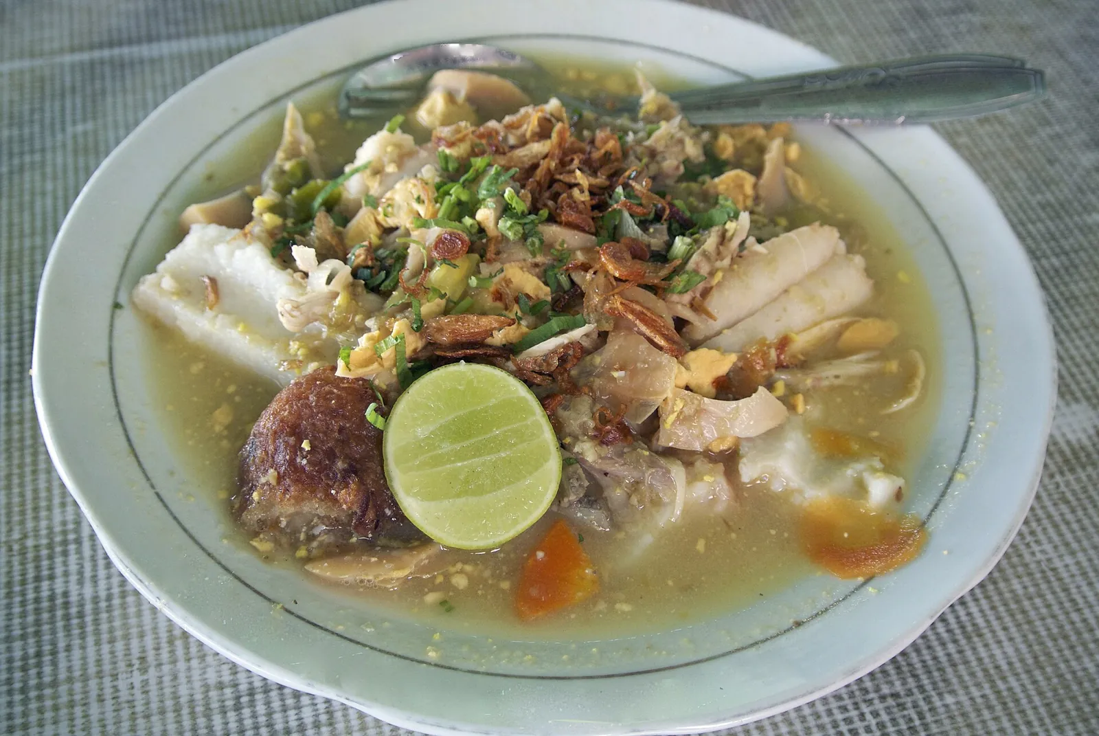 A bowl of soto Banjar with shredded chicken, rice cake, egg, fried shallots and a lime half