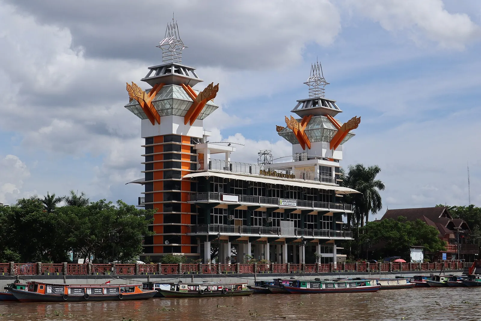 The orange and black Menara Pandang observation tower with carved roof ornaments beside the Martapura River, with boats moored below