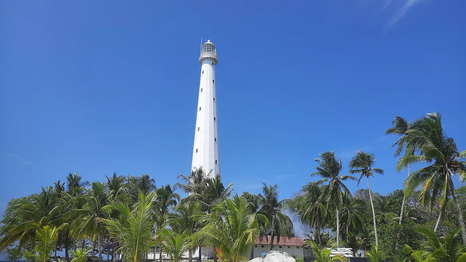 The white iron lighthouse on Lengkuas Island, Belitung, rising above coconut palms against a blue sky