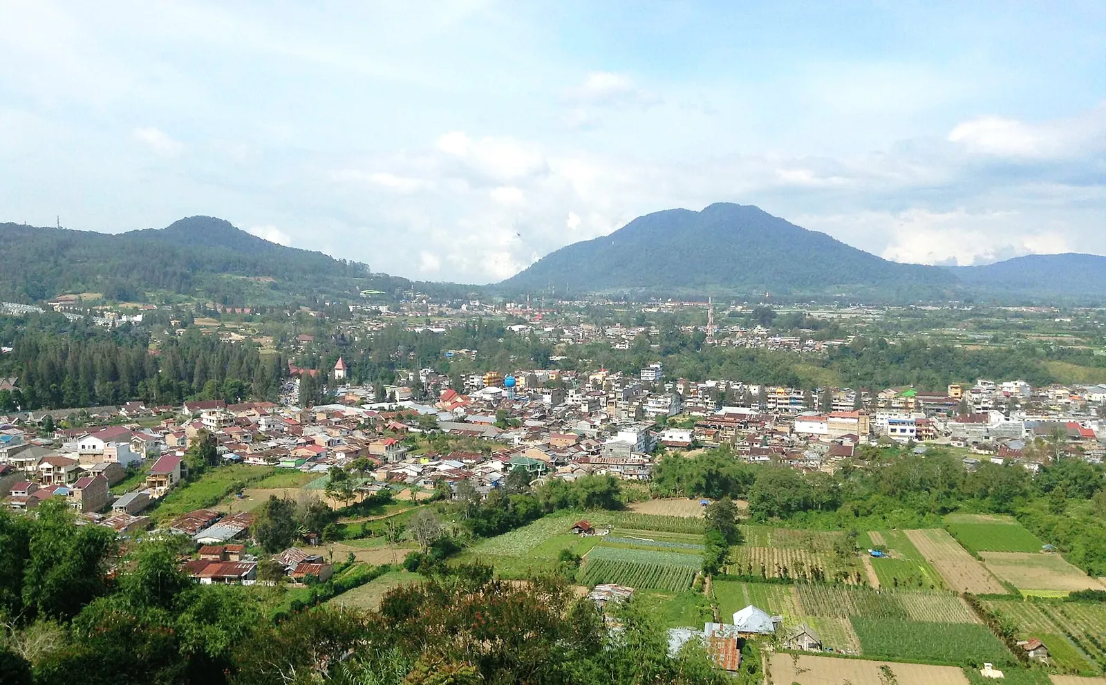 Berastagi's rooftops and vegetable fields seen from Gundaling Hill, with forested mountains under a blue sky