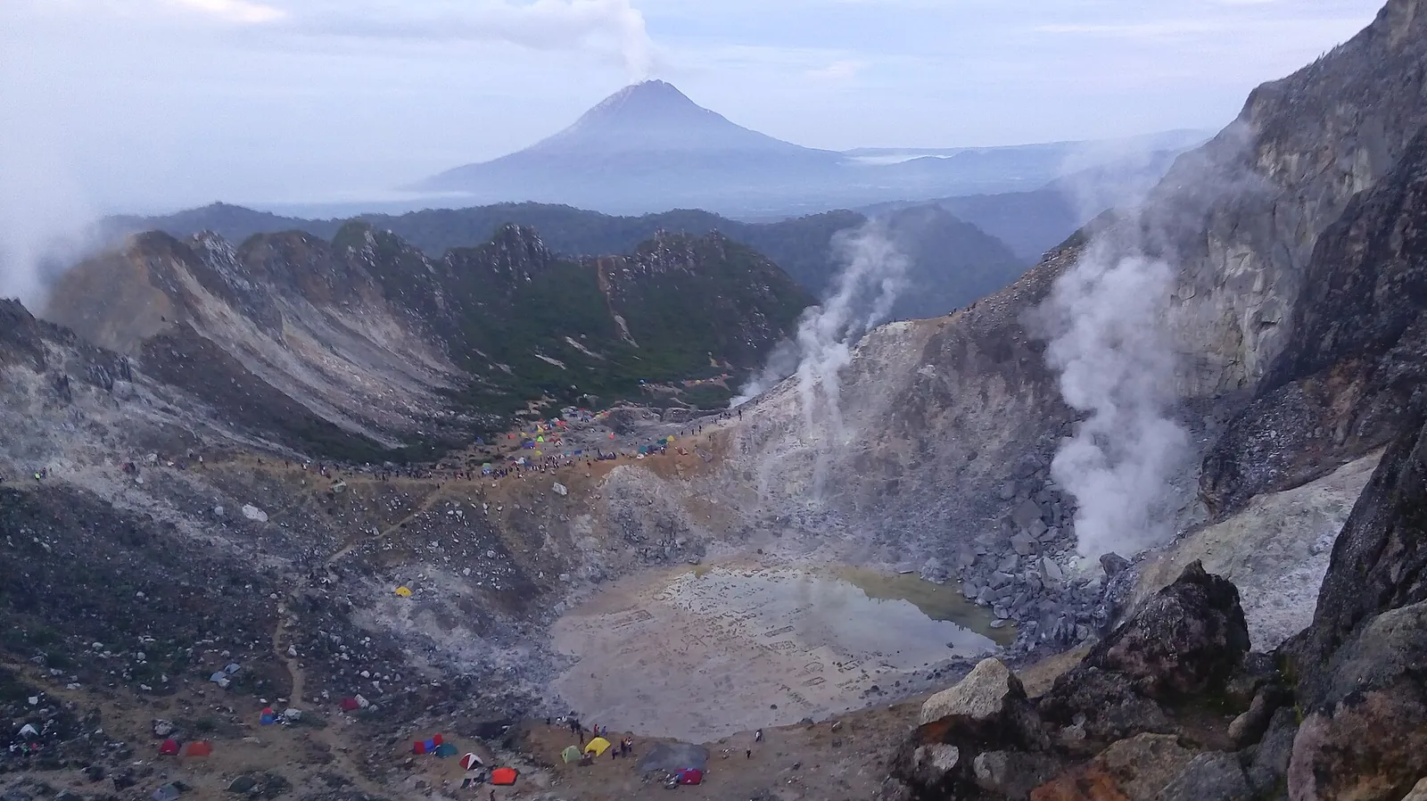 Steaming vents and the grey crater floor of Mount Sibayak, with the cone of Mount Sinabung in the distance