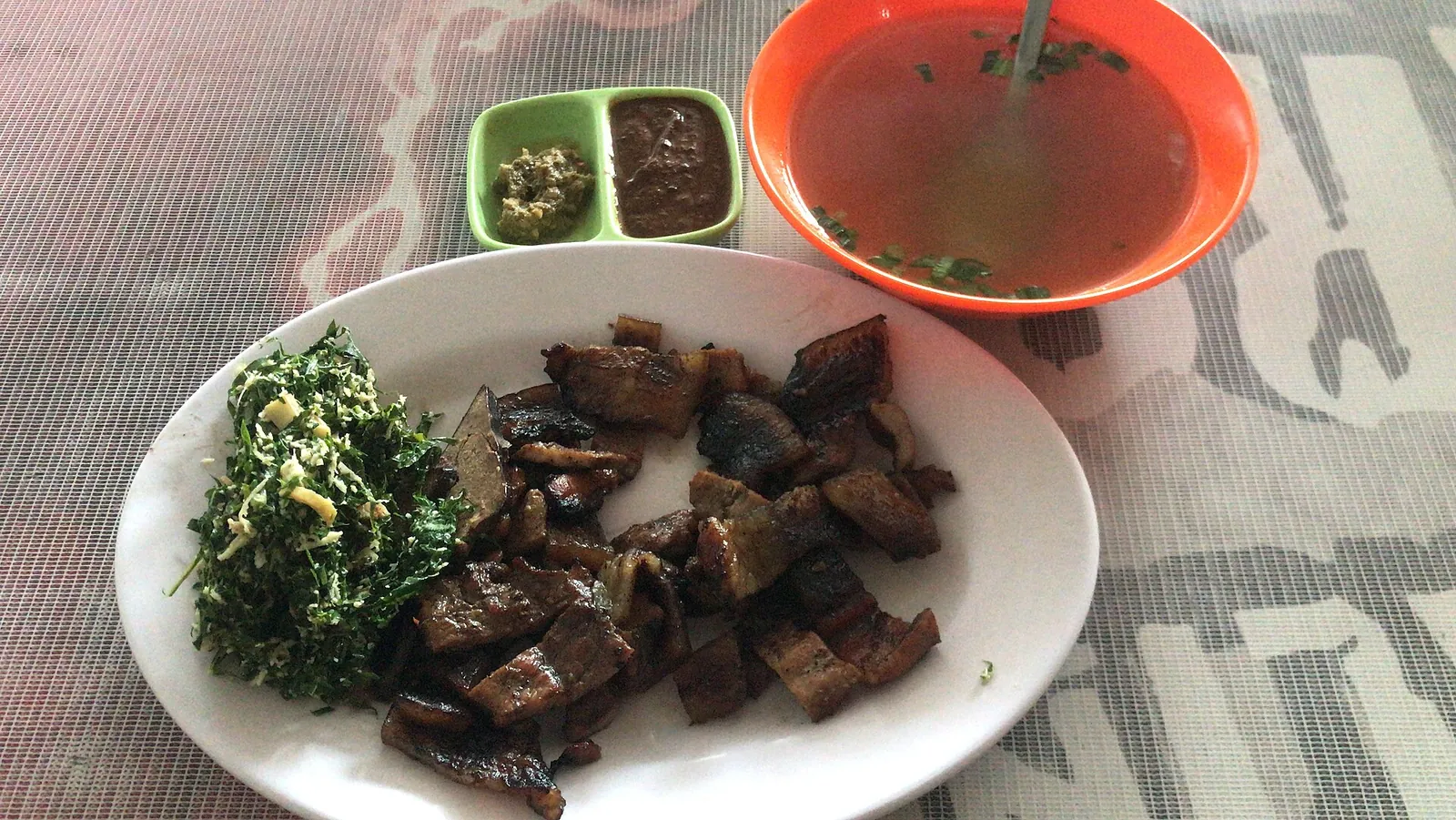 A plate of babi panggang Karo with grilled pork and cassava leaves, a bowl of broth and a small dish of dark sauce