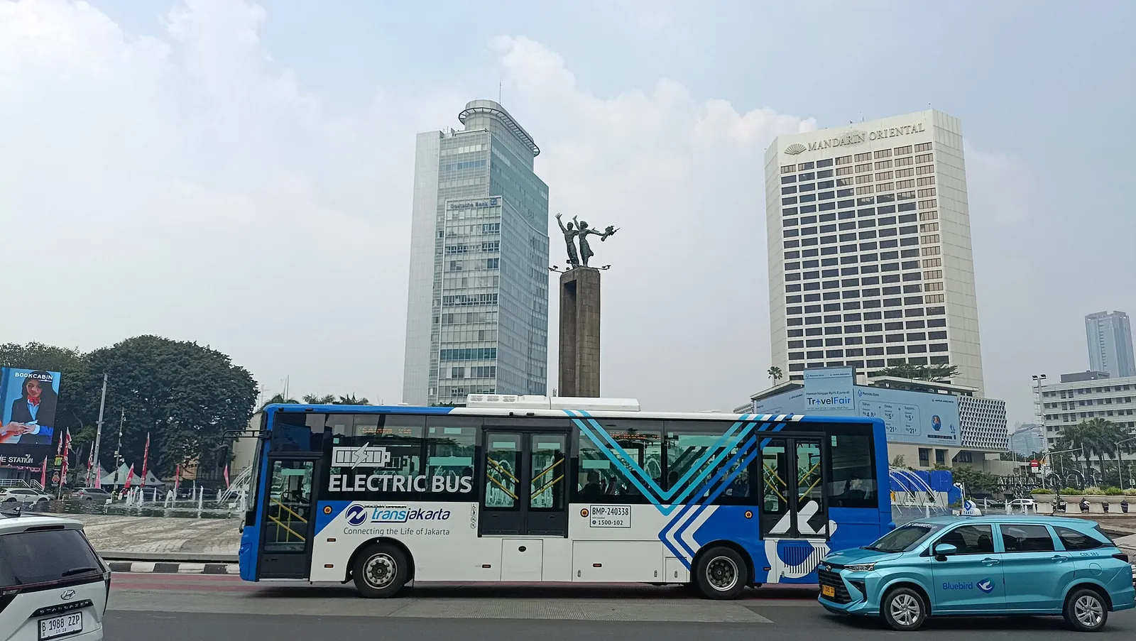 A blue and white Transjakarta electric bus passing the Welcome Monument at Bundaran HI in central Jakarta