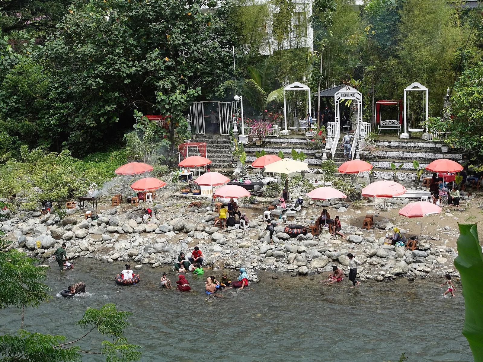 People swimming and floating on inner tubes in the Bahorok River below riverside parasols at Bukit Lawang