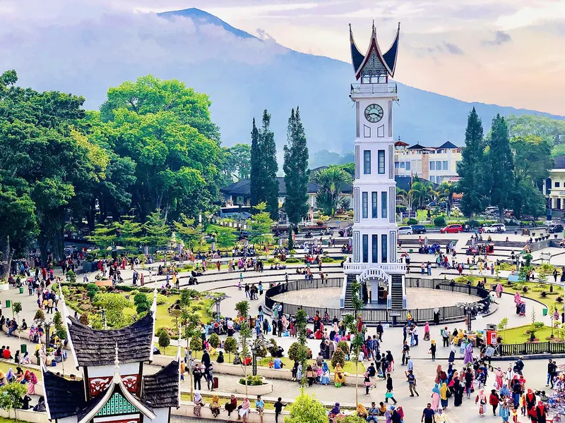 Jam Gadang clock tower with its horned Minangkabau roof above a crowded plaza in Bukittinggi, with a volcano behind