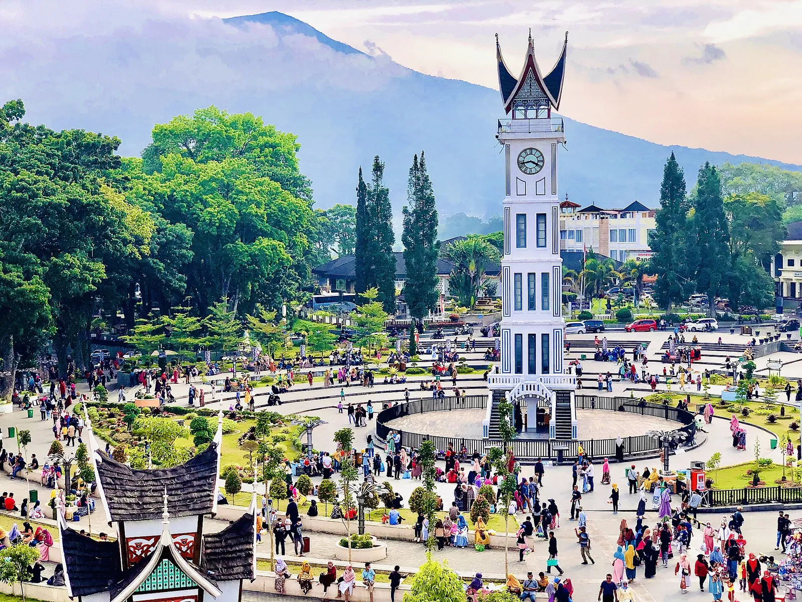 Jam Gadang clock tower with its horned Minangkabau roof above a crowded plaza in Bukittinggi, with a volcano behind