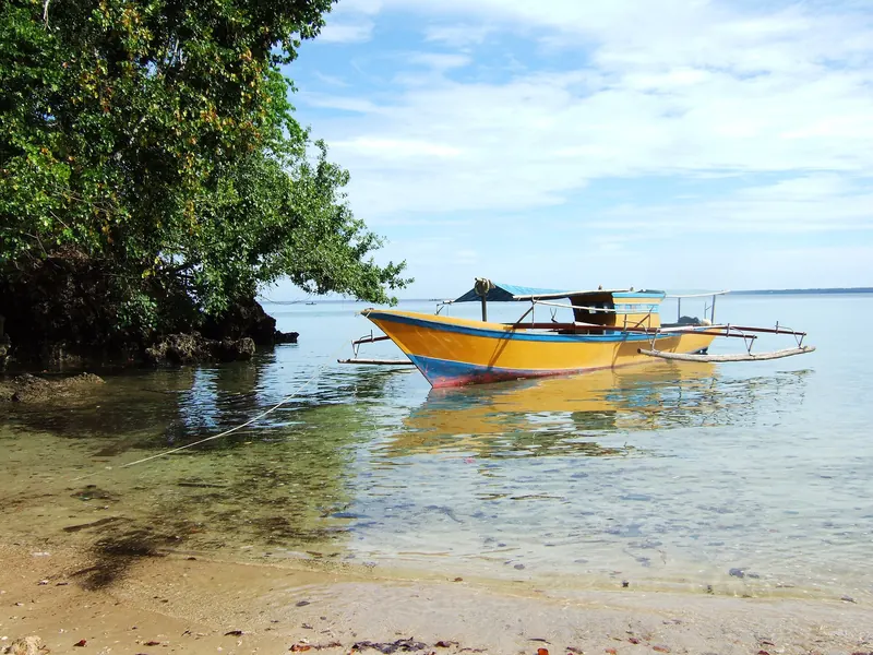 A painted wooden outrigger boat moored in clear shallow water beside a tree-covered rocky point at Tanjung Perigi, Bunaken Island