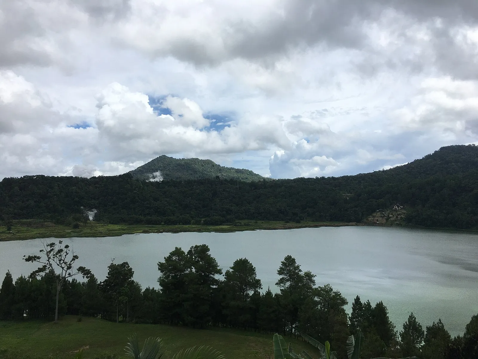 Lake Linow, a pale green volcanic lake ringed by pine forest, with forested hills under cloud behind