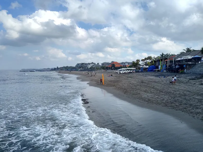 Waves running up a long grey-sand beach in Canggu, with beachfront cafés and umbrellas on the right