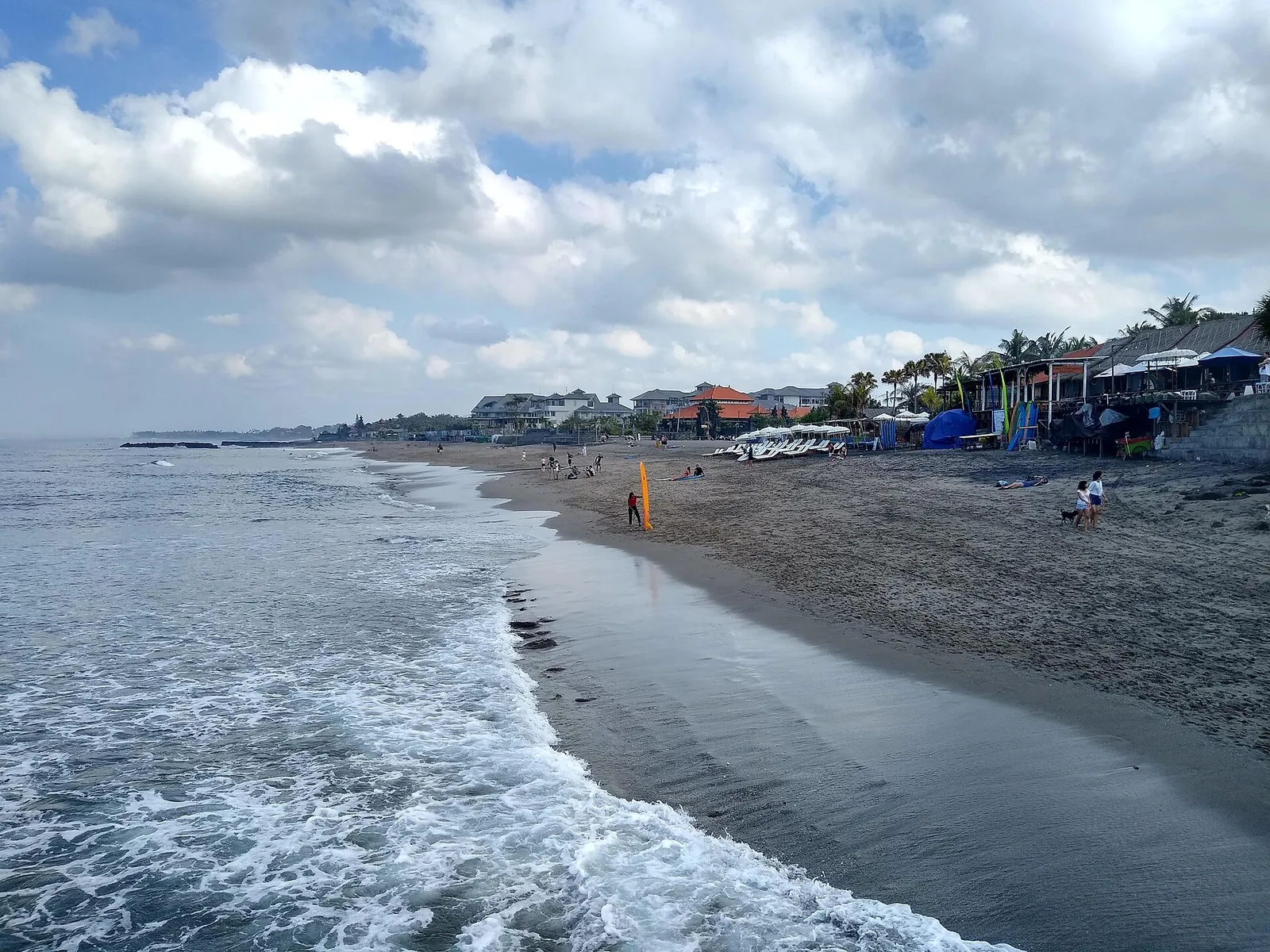 Waves running up a long grey-sand beach in Canggu, with beachfront cafés and umbrellas on the right