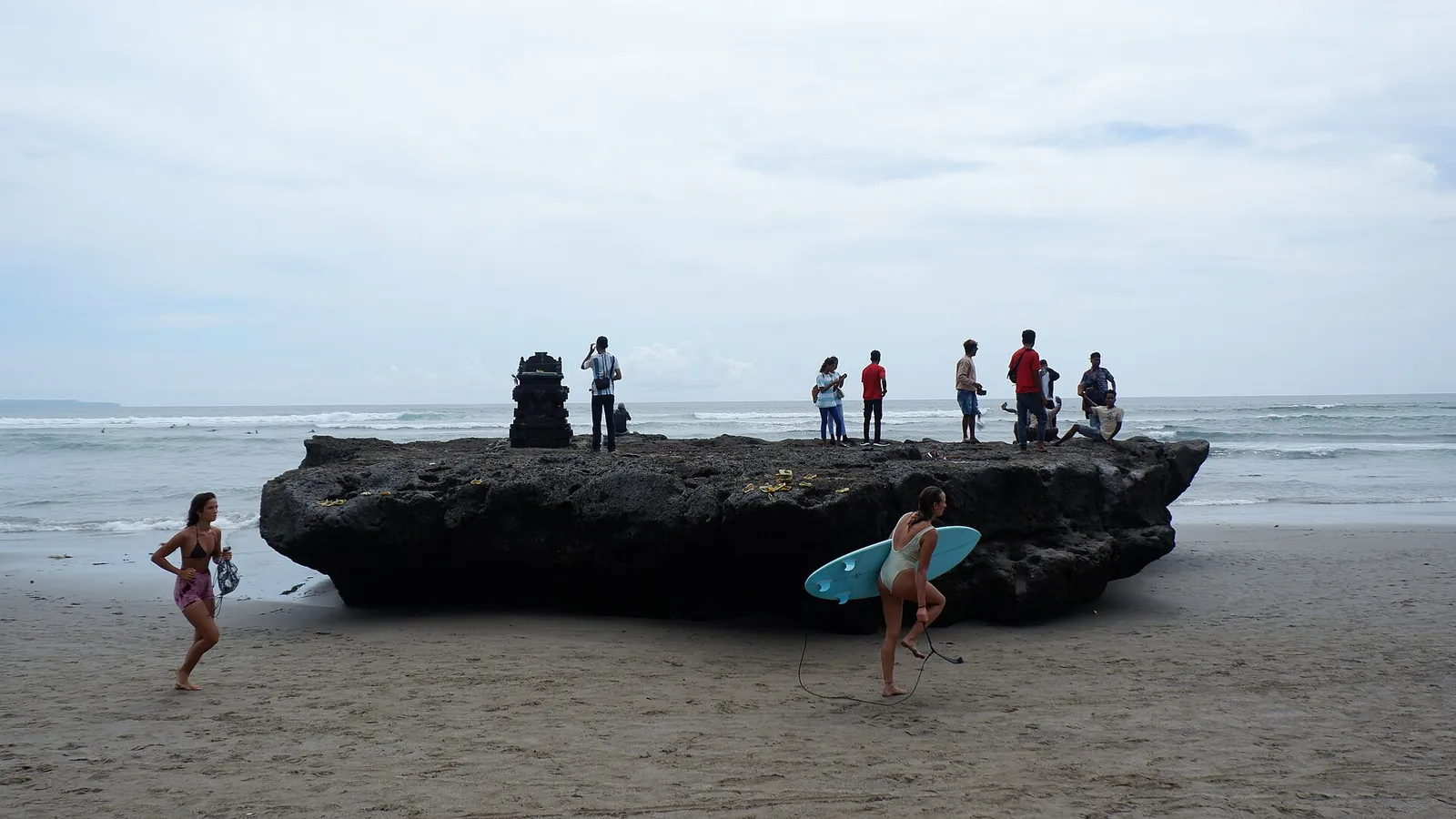 Surfers and visitors around a large black rock topped with a small shrine on Canggu Beach