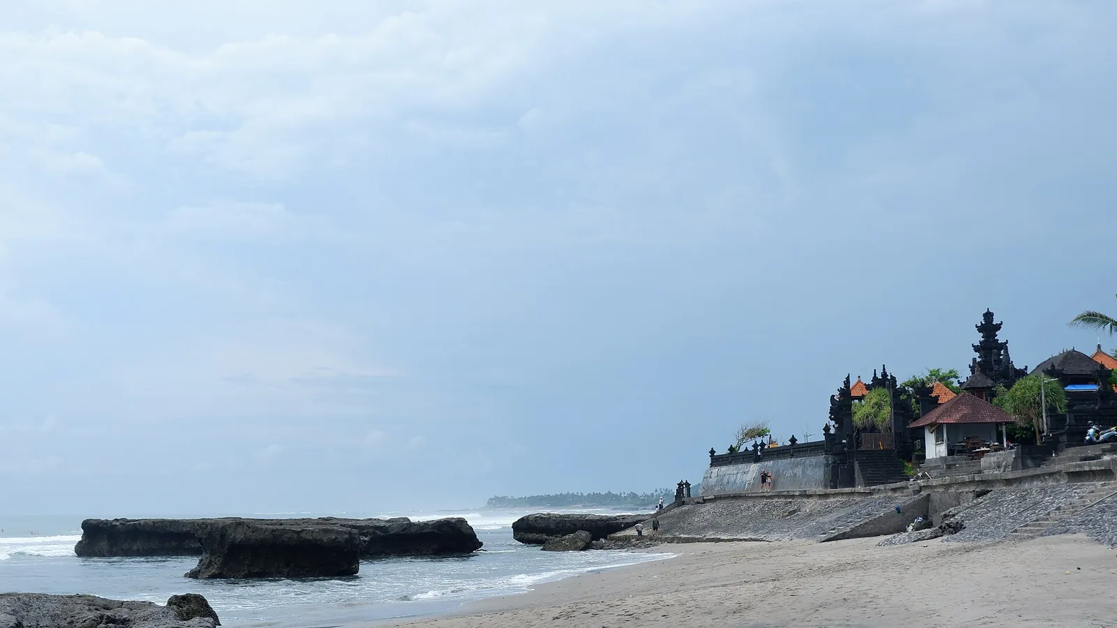 A Balinese temple with tiered black shrines on the shore at Canggu Beach, with flat dark rocks in the surf