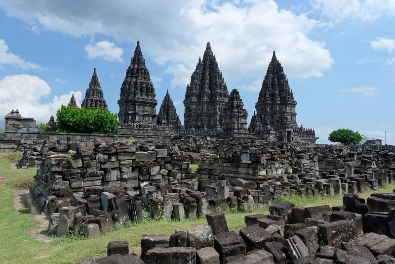 The towers of the Prambanan temple compound under a blue sky, with fallen stones of the ancillary shrines in the foreground