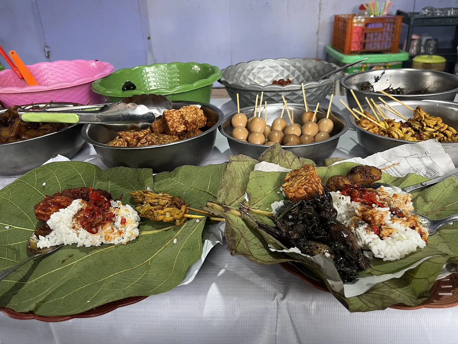 Portions of nasi jamblang rice on teak leaves with side dishes and satay skewers on a table