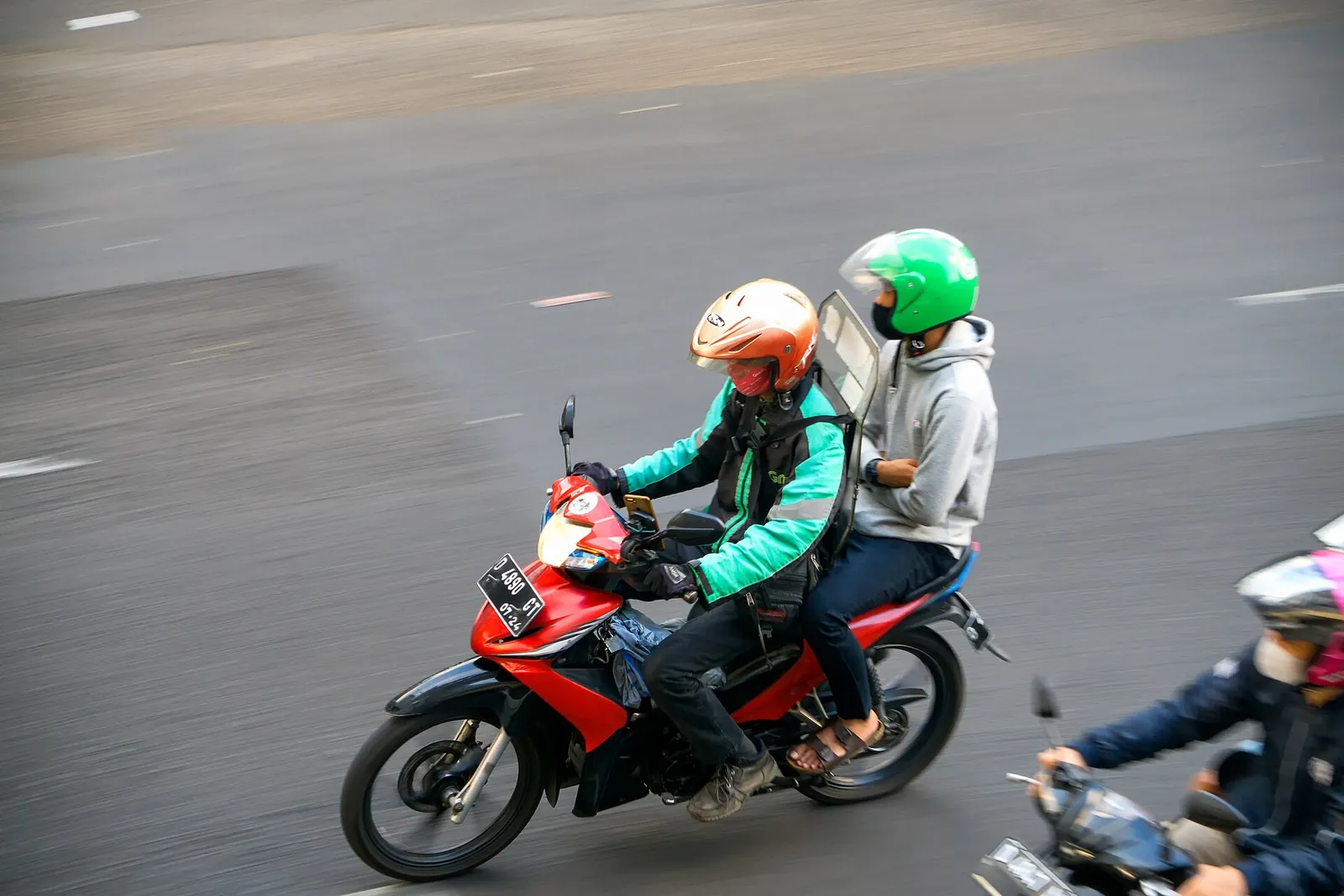 Ride-hailing motorbike driver in a green jacket carrying a passenger in a green helmet on a city road in Indonesia