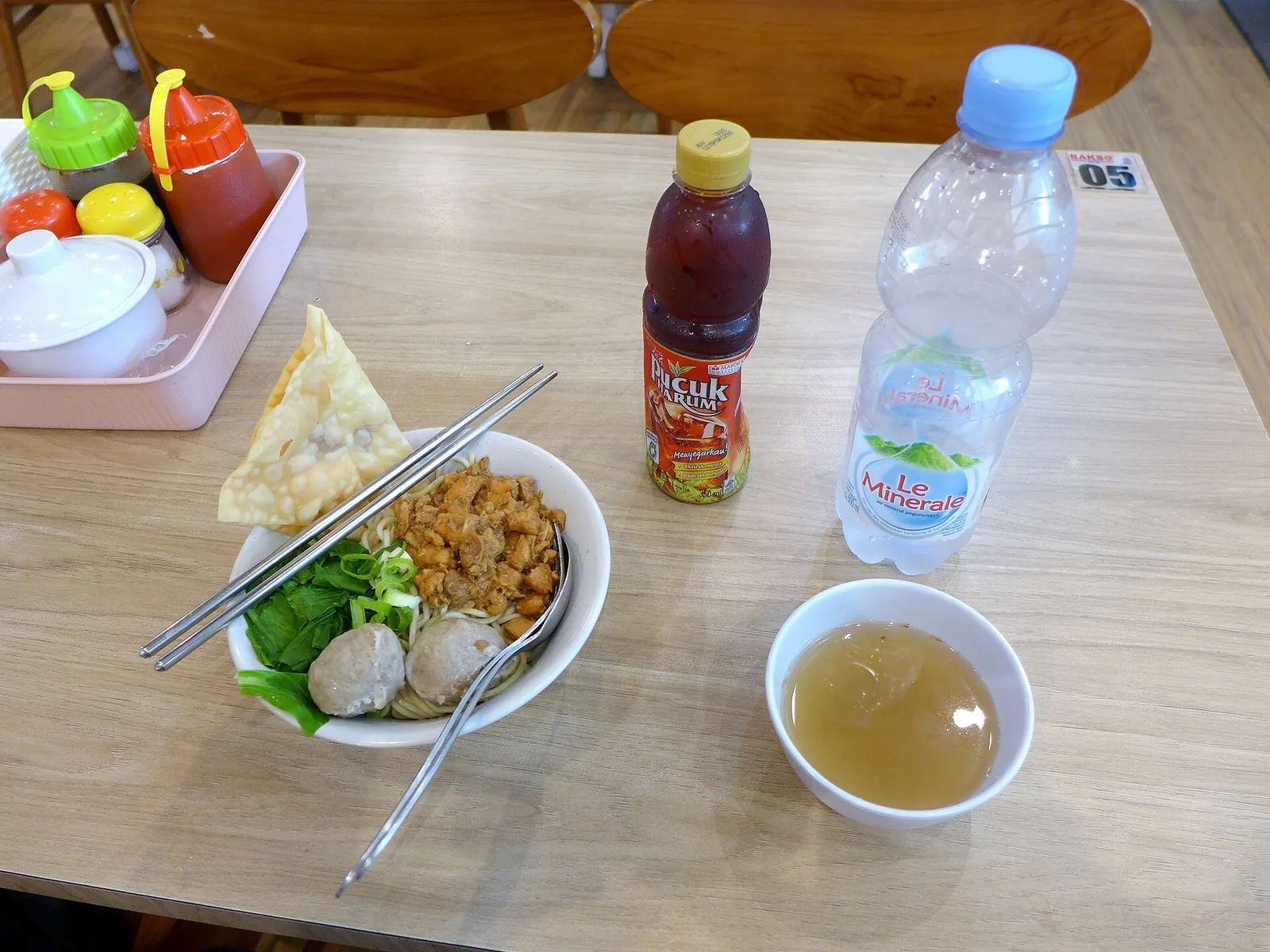 Bowl of mie bakso with meatballs and greens, a fried wonton, bottled tea and water on a table at Ngurah Rai Airport, Bali