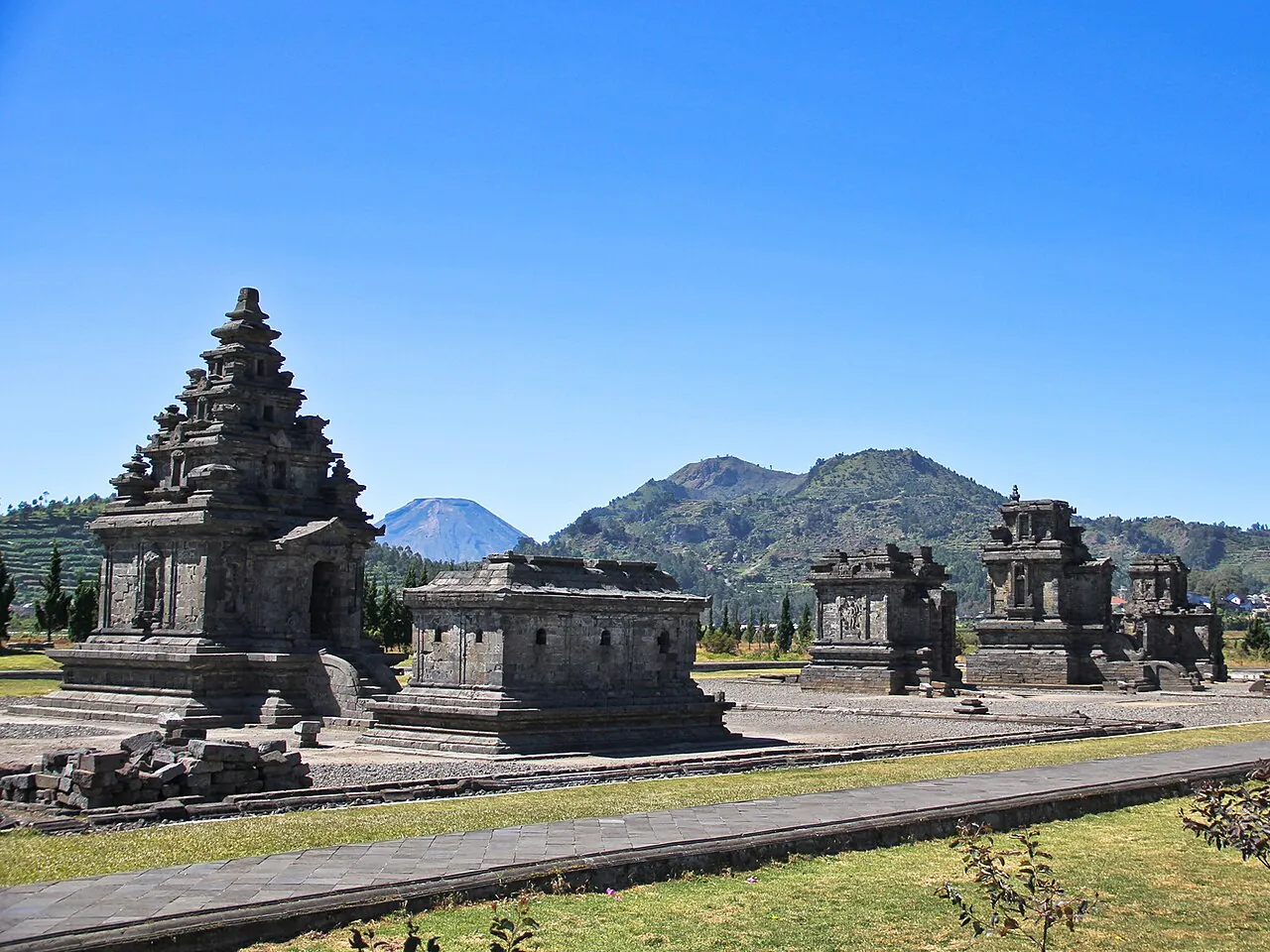 Stone Hindu shrines of the Arjuna temple complex on a lawn under a blue sky, with green hills behind, Dieng Plateau