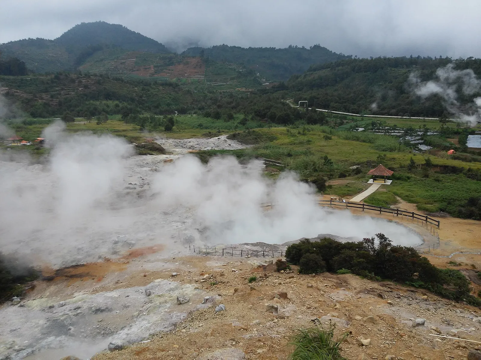 Steam rising from grey volcanic ground at Kawah Sikidang crater, with a fenced path and green hills, Dieng Plateau