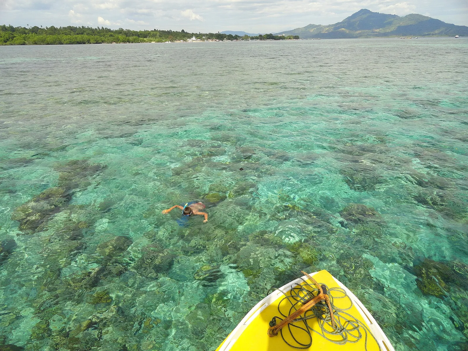 Snorkeller floating over a shallow reef flat in clear water off Bunaken, with a volcanic island on the horizon